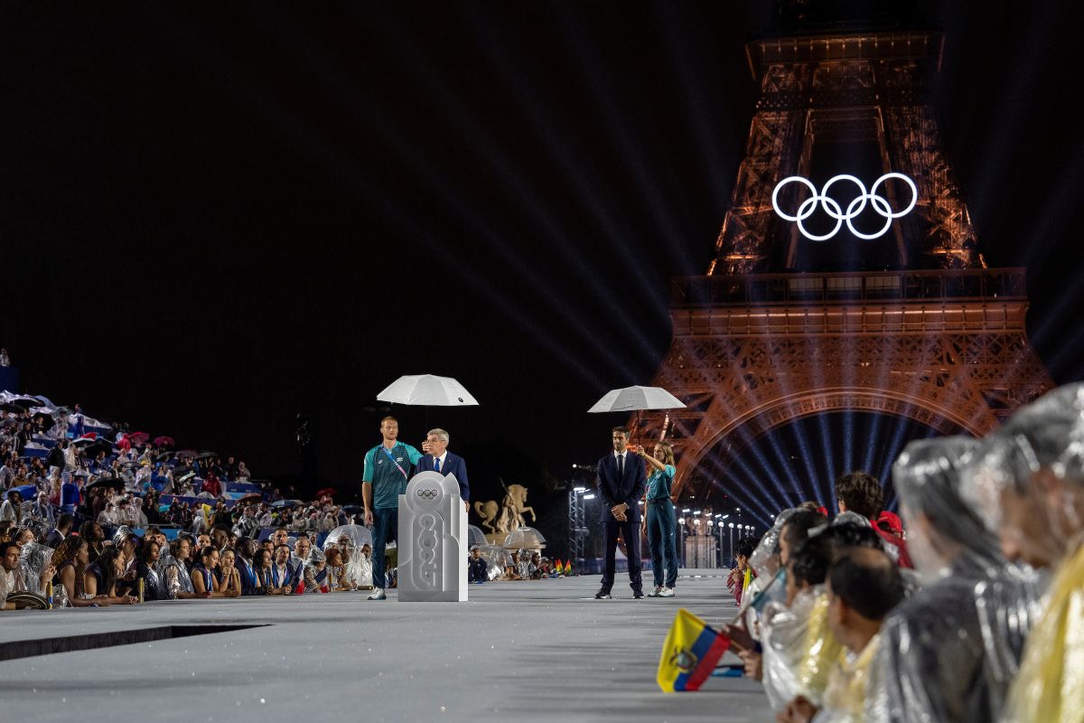 Nighttime event in front of the Eiffel Tower with speakers holding umbrellas on stage and audience in rain ponchos.