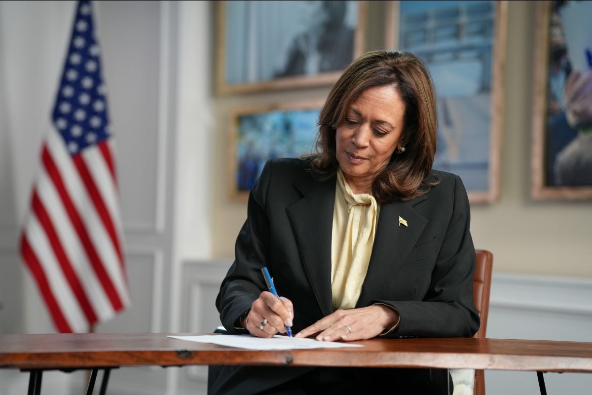 A woman in a blazer signing a document at a table with an American flag in the background.