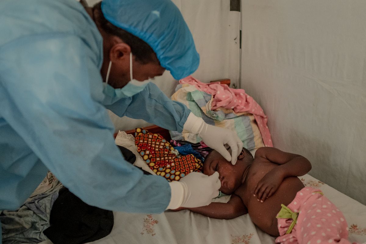 Healthcare professional in protective gear attending to a child lying on a bed.