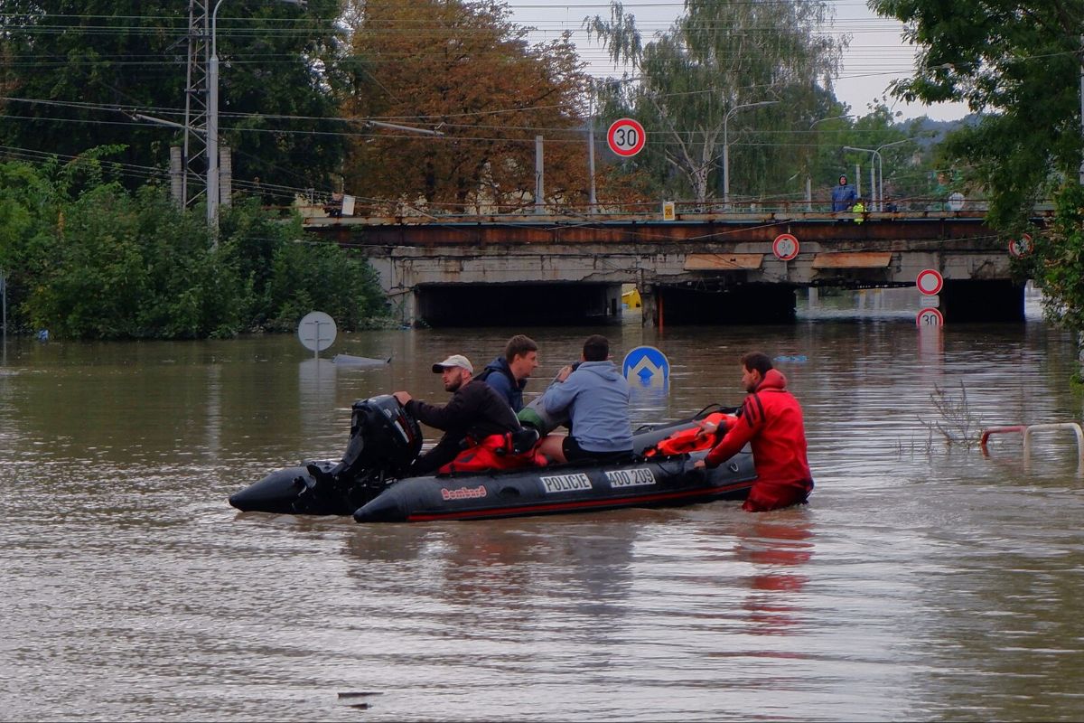 The image depicts a scene of urban flooding with four men navigating a partially submerged street in an inflatable rescue boat. The water level is high, covering more than half of the height of the road signs. Various traffic signs, some indicating speed limits and one-way directions, are partially submerged. In the background, there is a railway bridge with visible structural elements, such as beams and pillars, showing signs of rust and wear. Greenery, such as trees and bushes, surround the flooded area, and power lines are seen above the scene. The boat is labeled "POLICE," and the boat's motor and equipment are visible. One man, in a red outfit, is pushing the boat from behind, wading through the knee-deep water. The atmosphere appears overcast.