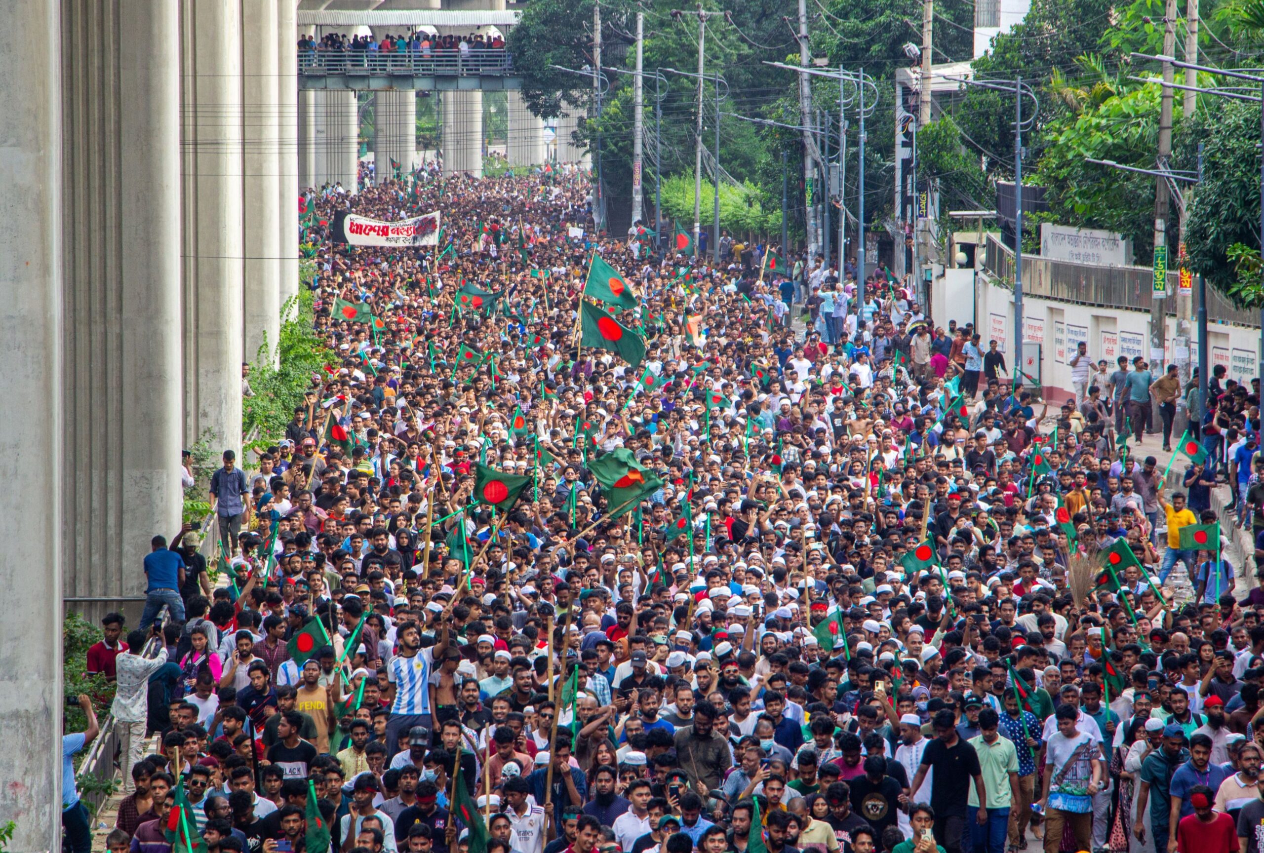 Large crowd in a street protest, many carrying Bangladeshi flags.