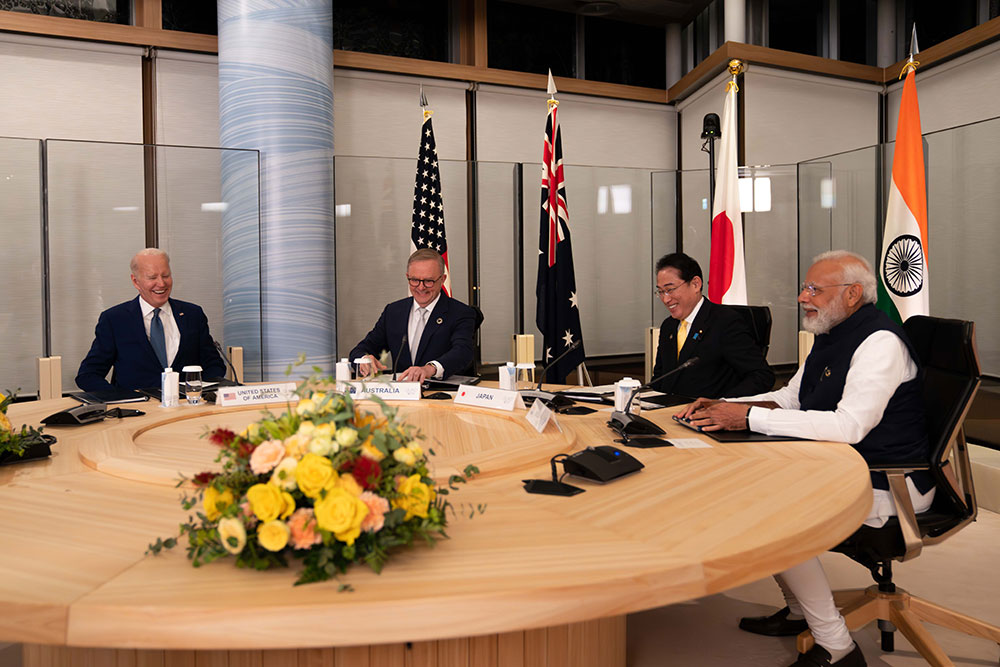 The image portrays a meeting of four individuals seated around a large, round wooden table. The table has a polished light wood finish and is adorned with a floral centerpiece featuring yellow, white, and red flowers with green foliage. Each person is seated in an office chair and positioned in front of a small nameplate. From left to right, the individuals are as follows: A man wearing a dark blue suit, white shirt, and tie, seated at the left end. A man dressed in a dark suit with a white shirt, seated next to the first man. Another man also dressed in a dark suit and white shirt, seated to the right of the second man. A man wearing a white shirt and a black vest, seated at the right end of the table. Behind the individuals are four flags from left to right: The flag of the United States. The flag of Australia. The flag of Japan. The flag of India. The backdrop includes a circular, light blue pillar and light-colored, frosted glass panels. The setting appears to be indoors, with wooden beams and alternating transparent and opaque window panes.