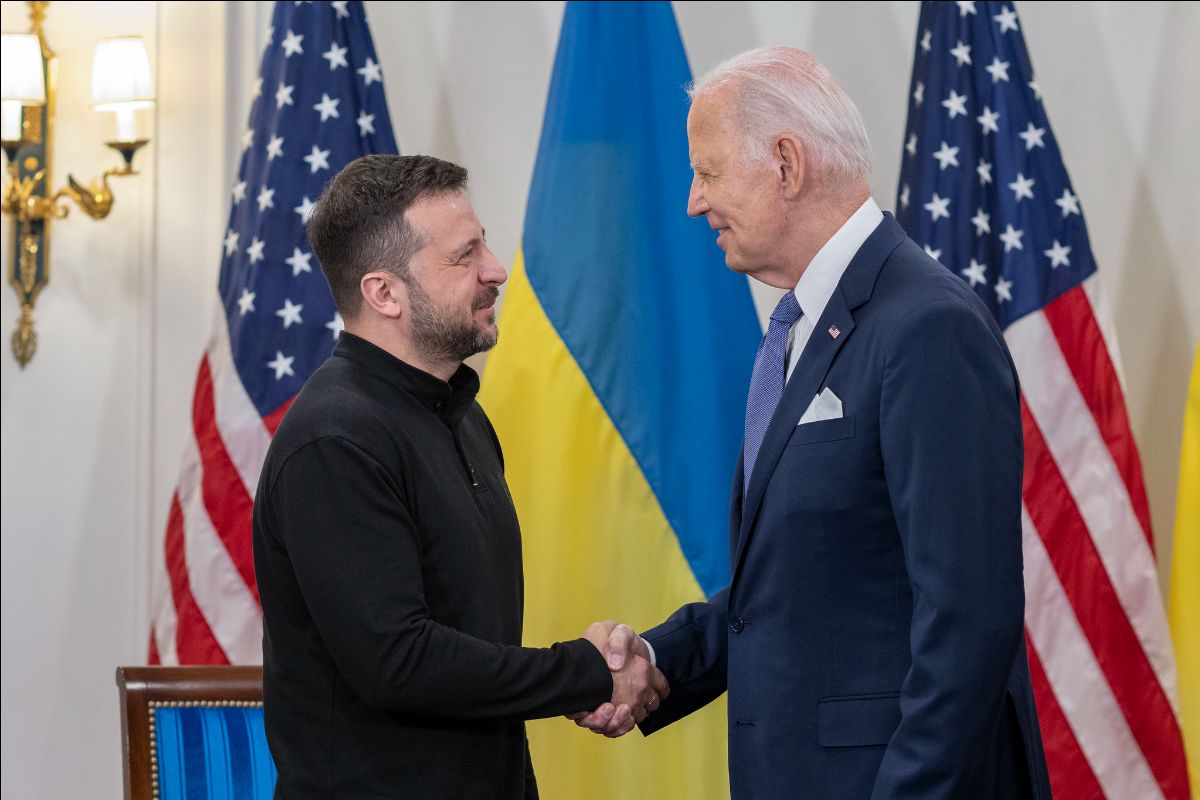 The image shows two men standing and shaking hands in front of a backdrop of two large American flags and two large Ukrainian flags. The man on the left is wearing a black long-sleeved top, while the man on the right is dressed in a blue suit with a white pocket square, white dress shirt, and blue tie. Both men appear to be smiling as they engage in the handshake. Behind them, part of a room is visible, including a wall-mounted light fixture with a white lampshade and gold accents. A blue chair with a wooden frame is partly visible on the left.