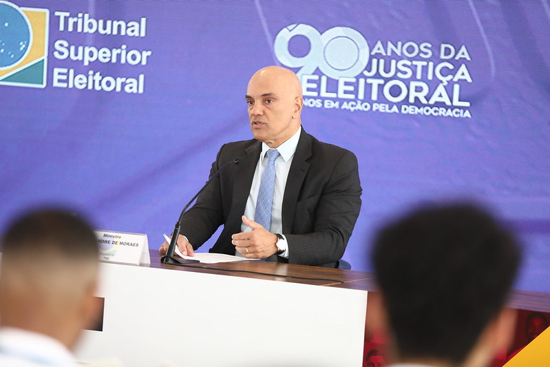 The image shows a formal event where a man, presumably a speaker or official, is seated at a table with a microphone. The man is dressed in a dark suit, white shirt, and light blue tie. He has a bald head and is gesturing with his hands while speaking. In front of him on the table, there is a nameplate that reads "Ministro Alexandre de Moraes" along with a smaller text "Presidente." The backdrop features a large, dark blue banner with logos and text related to a judicial electoral institution. The left side displays a logo with "Tribunal Superior Eleitoral," while the right side has a logo with the number "90" and the text "Anos da Justiça Eleitoral" followed by "90 Anos em Ação Pela Democracia." The image includes the blurred heads of attendees in the foreground.