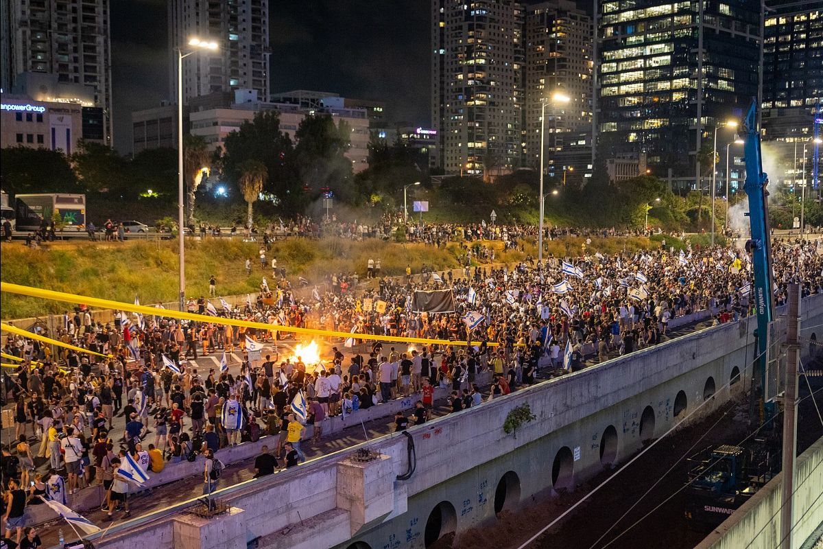 The image captures a large-scale outdoor protest during the night. The scene is densely packed with people, with the crowd spanning both a raised section and a road below. Many of the protesters are waving flags, some of which appear to be national flags, and holding signs. There are street lights illuminating the area, along with the lights from surrounding high-rise buildings and office complexes in the background. A small fire is burning in the middle of the road, surrounded by people. Additionally, there are yellow construction tapes running horizontally across the scene. The area is lined with greenery and trees, separating the elevated section from the lower roadway. The cityscape is visible in the background with tall buildings, some with illuminated windows.