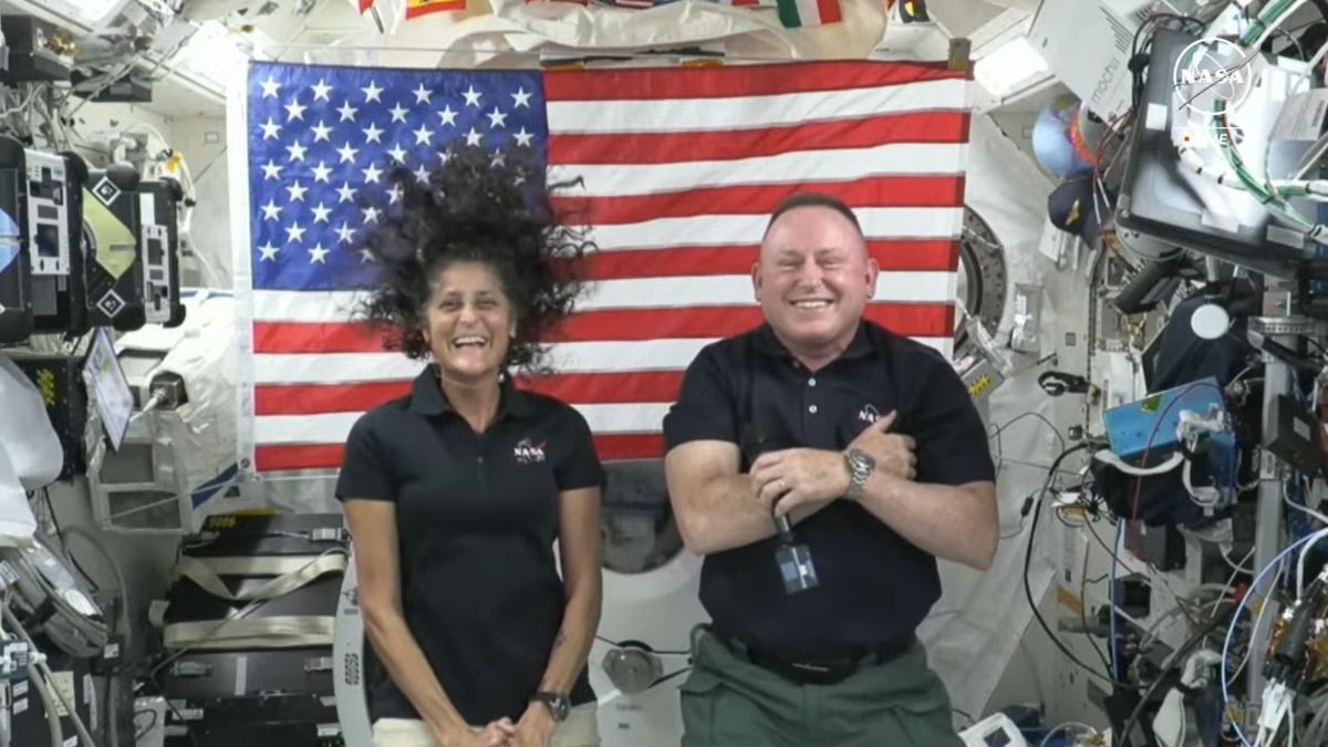 The image depicts two astronauts inside an International Space Station module. They are standing in front of a large American flag, which is prominently displayed on the back wall. The astronaut on the left has long, curly hair that is floating upwards, indicating zero gravity, and is wearing a black polo shirt with a NASA logo. The astronaut on the right, who has short hair, is also wearing a black NASA polo shirt and is smiling broadly with arms crossed, holding a microphone. The surrounding area is filled with various pieces of equipment, wires, and monitors typically found in the ISS. The lighting is bright, providing a clear view of the background details such as cables and labels attached to the equipment.