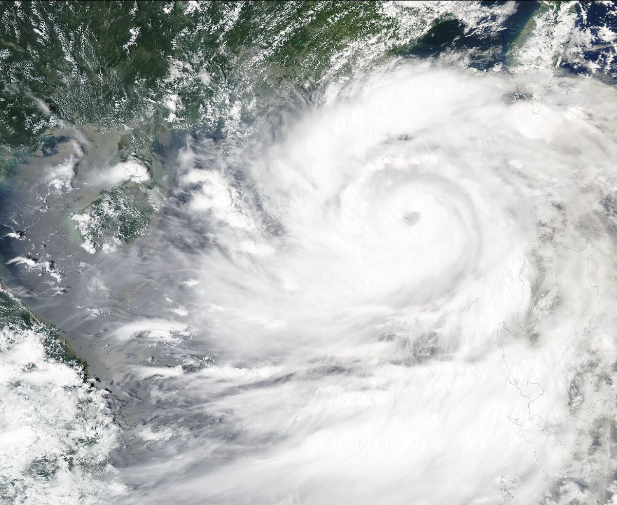 The image is a satellite view of a large, swirling cyclone over a landmass and the ocean. The cyclone's eye is nearly in the center of the image, characterized by a distinct, circular, cloud-free area. Surrounding the eye are dense bands of clouds radiating outward in a spiral pattern. The clouds appear white and thick, indicating a powerful storm. The landmass on the left side of the image is covered in a mix of green terrain and lighter patches, likely clouds or fog. The ocean near the land appears dark and murky, likely churned by the cyclone.