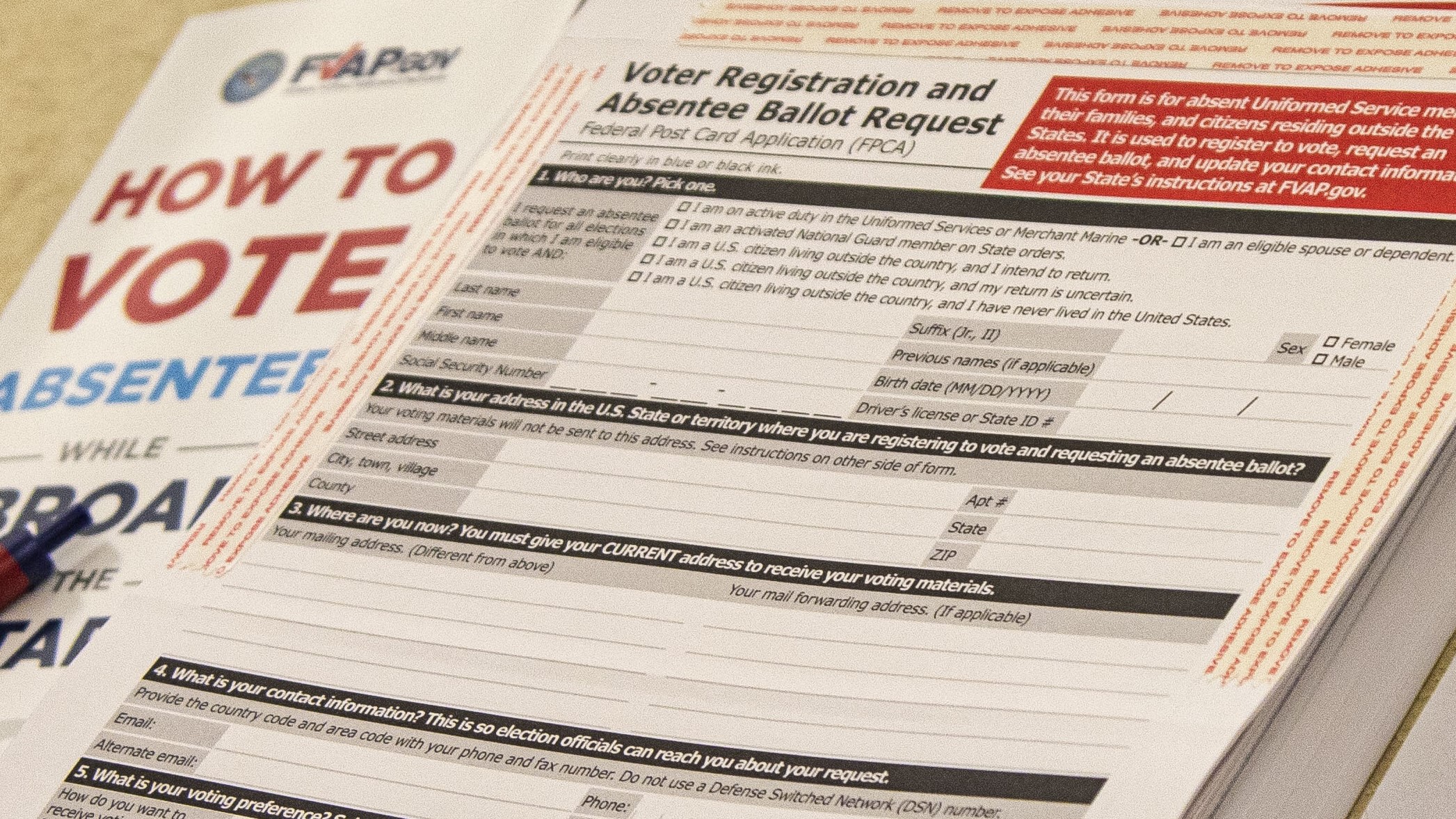 The image shows a close-up view of a Voter Registration and Absentee Ballot Request form, specifically the Federal Post Card Application (FPCA) form. The form is predominantly white with black and red text. The form contains various sections for personal information such as name, address, social security number, and contact information. To the left of the ballot request form, a section of a brochure with the text "HOW TO VOTE ABSENTEE WHILE ABROAD" in large, bold text is partially visible. The brochure has blue and red elements and is slightly blurred as it goes out of the focal plane. A red and blue pen is also partially visible near the left edge of the image. The background is mildly blurred, focusing attention mainly on the forms in the center.