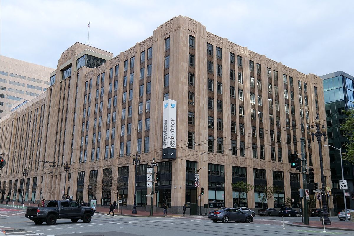 This image portrays a large, multi-story commercial building located at a street intersection in an urban area. The building is designed in an Art Deco style with a light brown, stone facade and evenly spaced vertical columns of windows. Prominently displayed on the corner of the building is a vertical sign with the "Twitter" logo and the Twitter handle "@twitter", each accompanied by the company's logo, a small blue bird. The ground level of the building features large windows with visible storefronts or office spaces. In the foreground, there is a city street with crosswalks, traffic lights, and various vehicles, including cars and a black pickup truck, as well as a few pedestrians. Street signs indicating a speed limit and directions are also visible, along with street lamps and overhead utility wires. The sky above is partly cloudy.