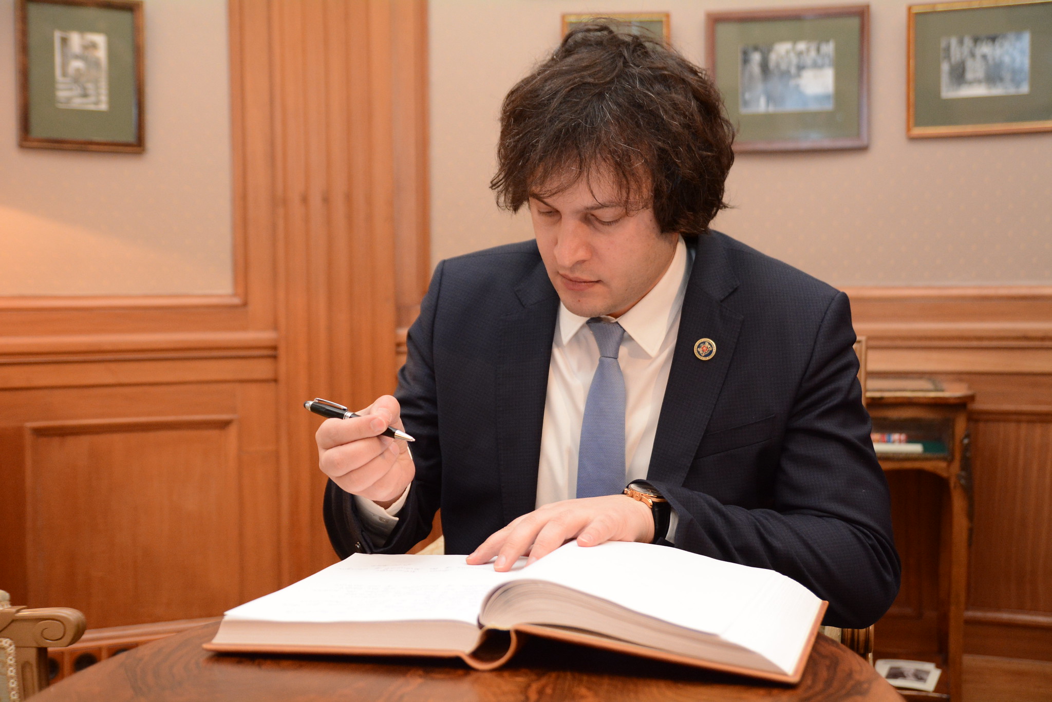 The image shows a person seated at a wooden table, writing in a large open book with a pen in hand. The individual is wearing a dark blue suit, a light blue tie, and a watch on their left wrist. The room's decor features wooden panels and bookshelves, with several framed, abstract artworks hanging on the wallpapered walls. The lighting is warm, casting soft shadows around the room.