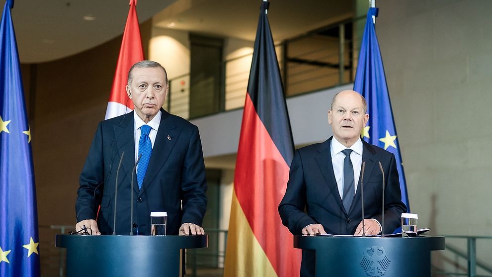 The image shows two men standing behind podiums with microphones in a formal setting. The man on the left is dressed in a dark suit with a white shirt and a blue tie. The man on the right is also in a dark suit with a white shirt and a dark tie. Between them is the German flag, and on either side are the European Union and Turkish flags. The background features a modern architectural setting with clean lines, a staircase, and soft lighting. Each podium has a glass of water placed on it.