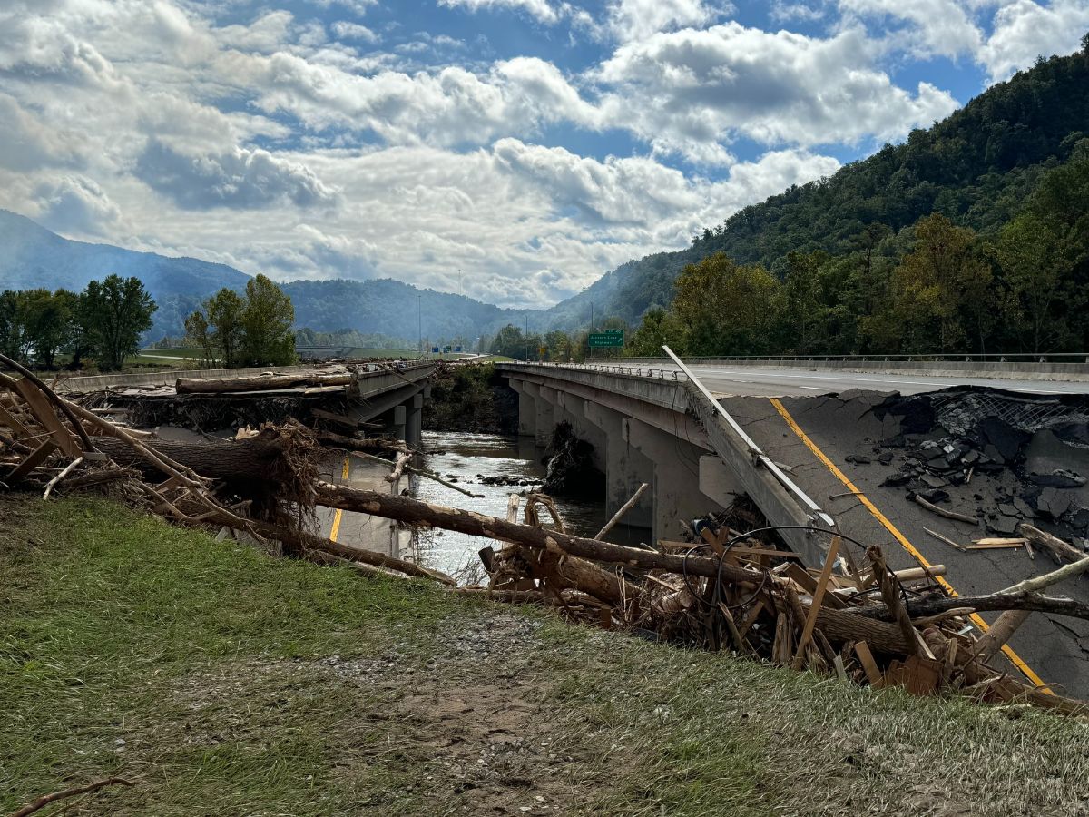 The image depicts a partially collapsed bridge over a river. The center section of the bridge is missing, revealing debris and broken tree trunks scattered across the structure. The remaining parts of the bridge are visibly damaged, with large cracks and sections of asphalt peeling away. Surrounding the bridge, lush green hills are visible under a partly cloudy sky, enhancing the sense of natural landscape disturbed by the bridge's damage.