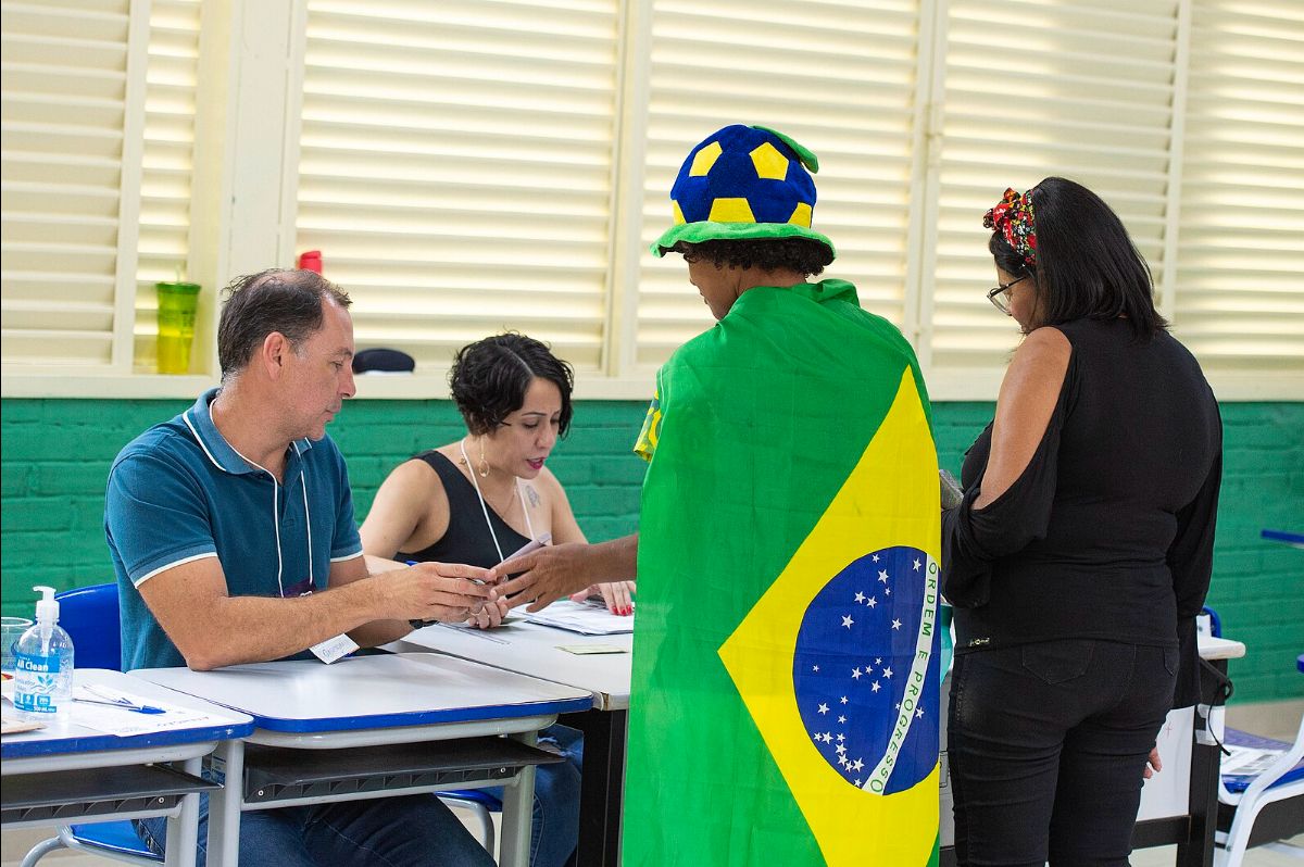 The image depicts an indoor scene with four people engaged in an interaction, possibly in a voting or registration setting. Two people are seated at a white desk with papers and a pen. The person on the left is wearing a blue polo shirt and a name tag, holding an object. The person in the center has short curly hair, wears a black sleeveless top, and is focused on the papers. Standing in front of them is a person draped in a Brazilian flag, wearing a hat shaped like a soccer ball with yellow and blue panels. Another person stands to the right, wearing a black outfit and glasses, observing the scene. The background shows green walls and white window blinds.