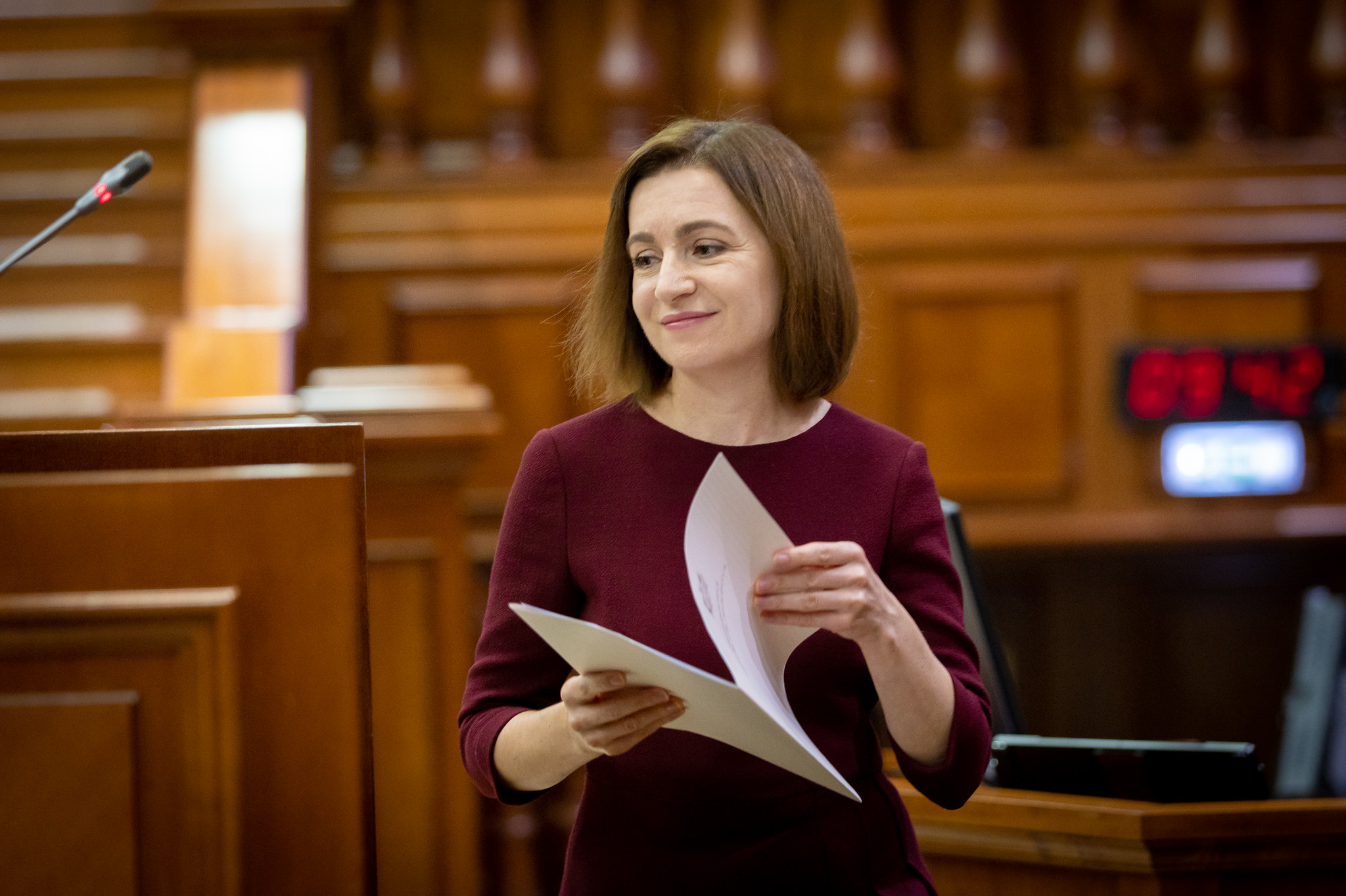 The image shows a person standing in a formal setting, likely a courtroom or assembly hall. They are wearing a burgundy dress and holding a stack of papers. The background features wooden paneling, creating a warm, formal environment. A microphone is visible on the left side, and a digital clock displaying "05:42" is in the background on the right.