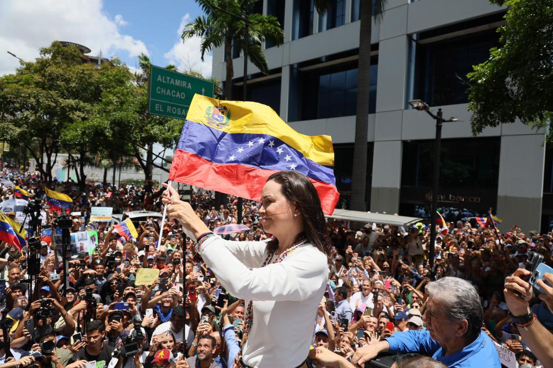 The image depicts a crowded public rally in an urban area featuring a large group of people gathered in an open space. A woman with long dark hair, dressed in a white top, stands on an elevated platform or vehicle and prominently waves a Venezuelan flag. The flag has three horizontal stripes: yellow at the top, blue in the middle with white stars, and red at the bottom. She appears to be engaging with the crowd, many of whom are holding cameras, phones, and smaller Venezuelan flags. The background includes lush green trees, a blue sky with some clouds, and a modern building with large windows. Overhead, a green traffic sign shows destinations: “ALTAMIRA,” “CHACAO,” and “EL ROSAL.”