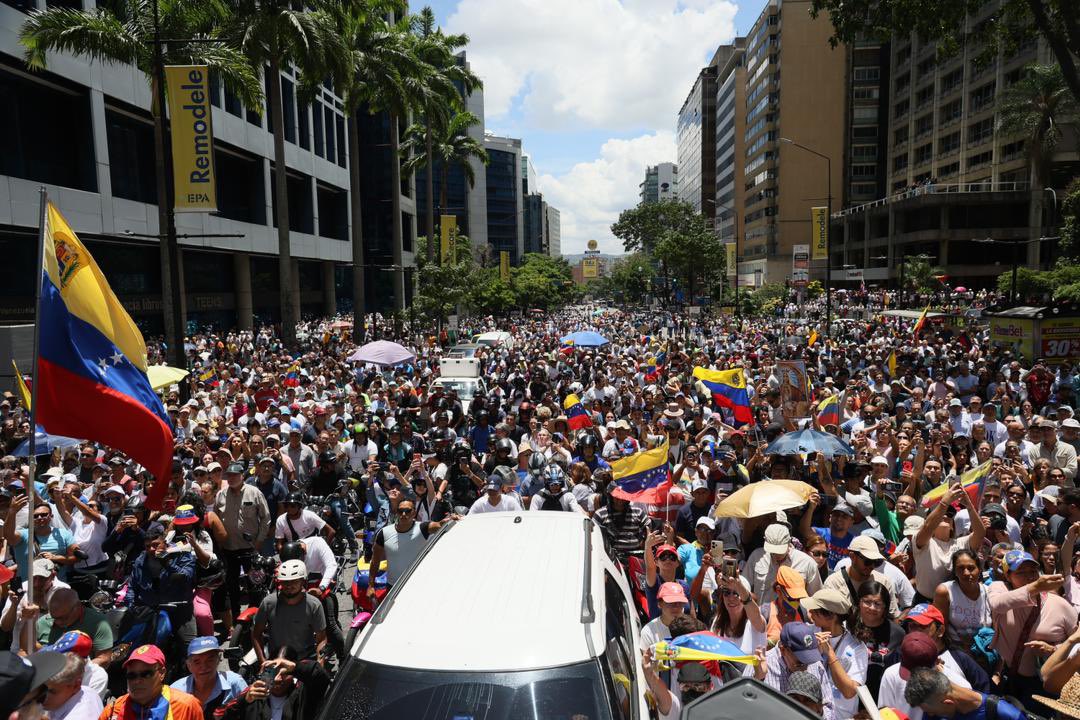 The image depicts a large-scale protest taking place on a city street densely packed with people. The crowd stretches far into the background, filling the street in front of tall office buildings and apartments. Many participants are holding Venezuelan flags, creating a sense of unity and national pride among the protesters. The flags are vibrant with horizontal stripes of yellow, blue, and red with stars. Among the crowd, some individuals are holding umbrellas to shield themselves from the sun, while others have cameras and mobile phones raised, capturing the event. The sky is mostly clear with a few scattered clouds, indicating a sunny day. Trees line one side of the street, offering some greenery amidst the urban environment.