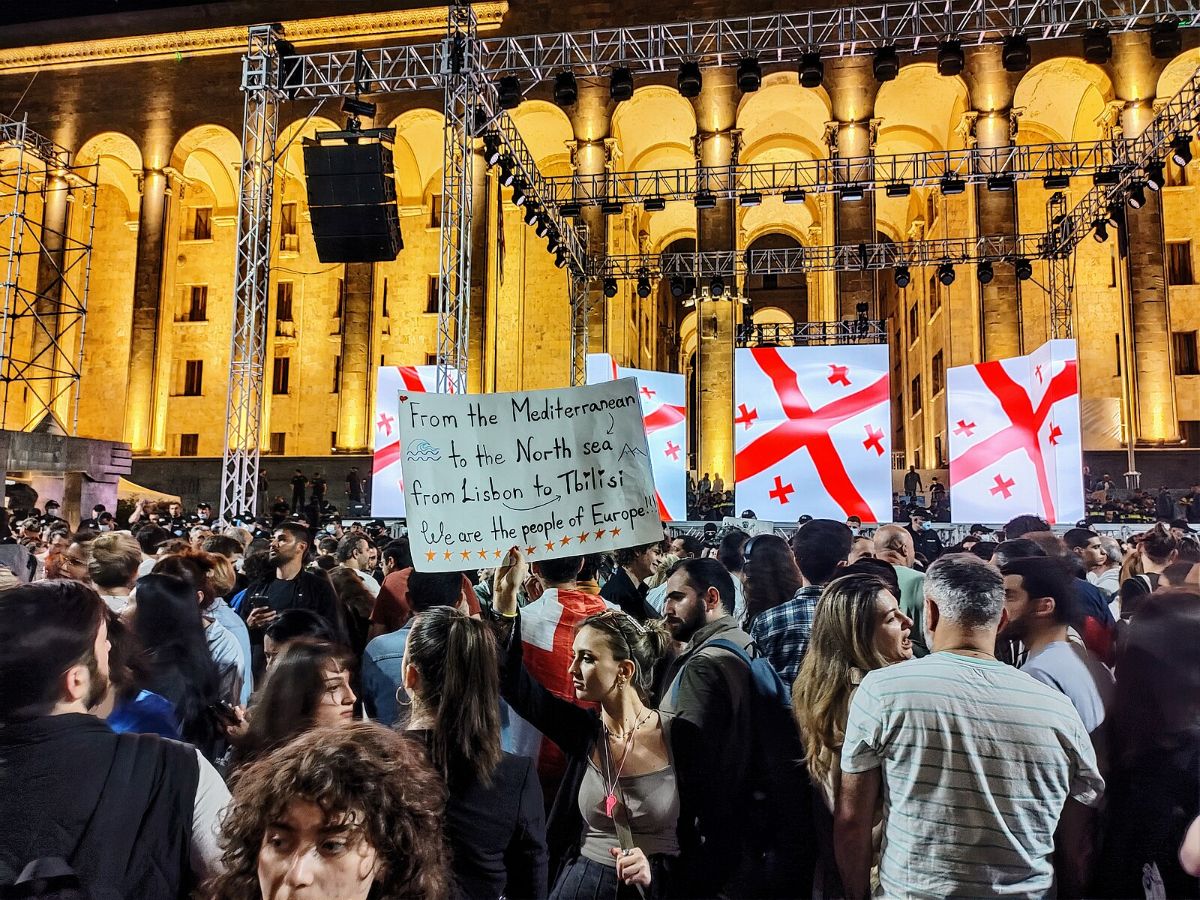 The image shows a large crowd gathered in front of a grand, illuminated building with tall, arched windows and columns. The building is lit with warm yellow lights. In the foreground, there are many people, some facing the stage and others engaged in conversation. In the middle of the crowd, a person holds up a sign with handwritten text. Behind the crowd, there is a stage with large screens displaying the flag of Georgia, featuring a bold red cross and four smaller crosses on a white background. Metal scaffolding and lighting fixtures are visible above the stage area.