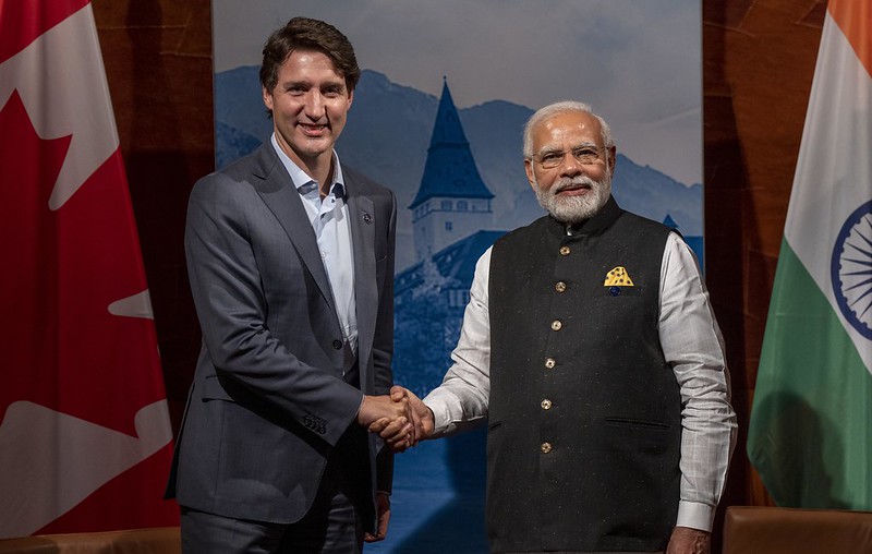 Two men are shaking hands in front of a backdrop depicting a scenic mountain landscape with a building featuring a conical roof. The man on the left is dressed in a dark gray suit with a white shirt, and the man on the right is wearing a black vest over a white shirt. They are standing in front of two flags: the Canadian flag on the left and the Indian flag on the right. The Canadian flag is red and white with a maple leaf, while the Indian flag features horizontal stripes of orange, white, and green with a blue Ashoka Chakra in the center.