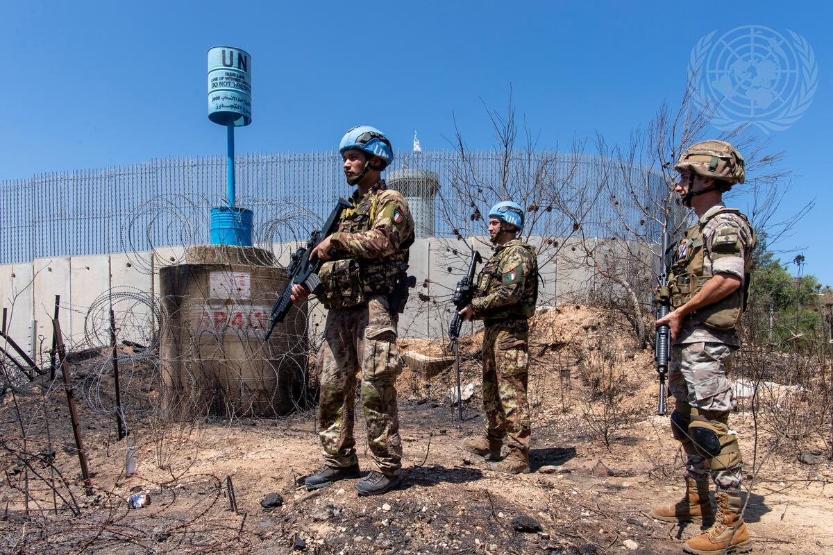 In this image, three soldiers wearing camouflage uniforms and blue helmets marked with "UN" are standing on a dry, rocky terrain. The soldiers are alert, holding rifles, and appear to be on guard. Each soldier has distinct patches on their uniforms. Behind them is a fence topped with barbed wire and a concrete structure. A blue cylindrical container marked with "UN" is elevated on a pole. The sky is clear and bright blue, indicating a sunny day. There are sparse, dried bushes around the soldiers, adding to the arid setting.