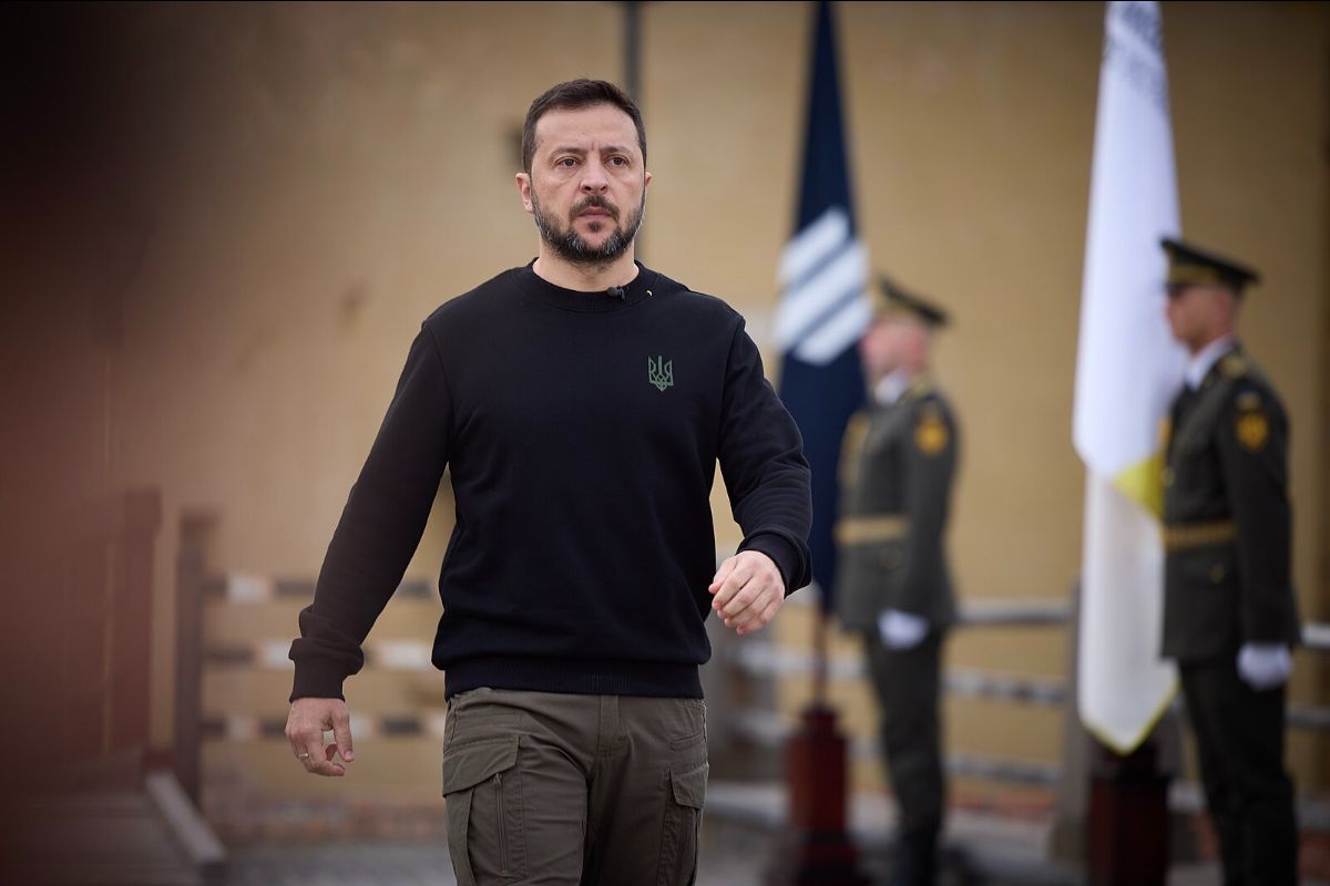 The image features a man with short hair and a beard, wearing a black long-sleeved shirt with a small emblem on the left side of the chest. He is also wearing olive green cargo pants. He is in focus and walking towards the camera with a determined expression. In the background, slightly out of focus, are two uniformed guards standing at attention. Behind them are two flags, one predominantly blue with a set of white diagonal stripes, and the other mainly white with a section of yellow.