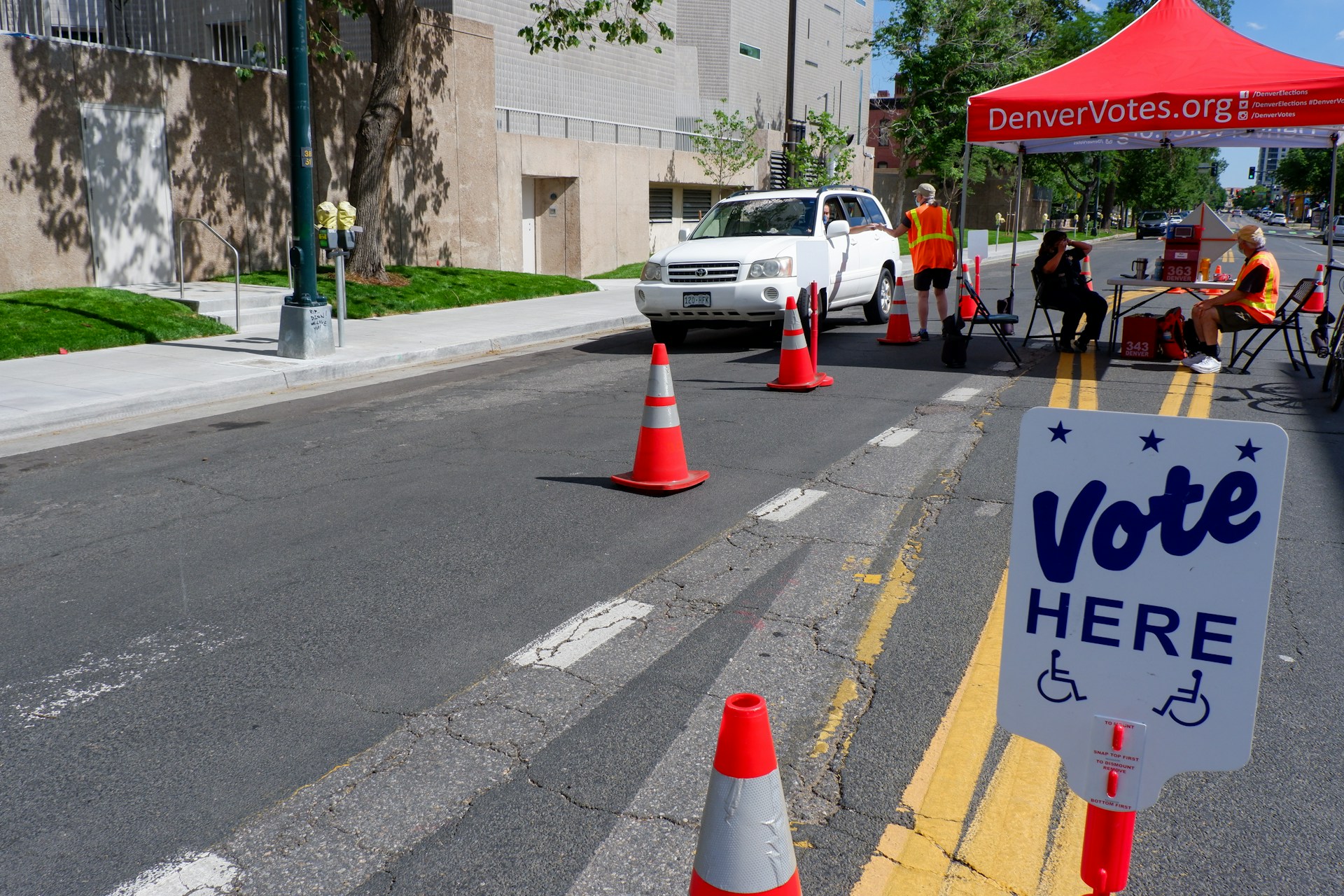 The image shows a drive-up voting station on a sunny day. A white SUV is stopped in the middle of the road, surrounded by orange traffic cones directing the vehicle through. On the right side, there is a red tent with the text "DenverVotes.org" printed on it. Two attendants wearing yellow reflective vests are visible; one is standing next to the vehicle's window, while the other is seated under the tent. A table with voting materials is set up beneath the tent, indicating this is a voting station. In the foreground, a sign marked "Vote Here" with icons indicating accessibility is prominently displayed. The background features a beige building with a tree and well-maintained grass along the sidewalk.