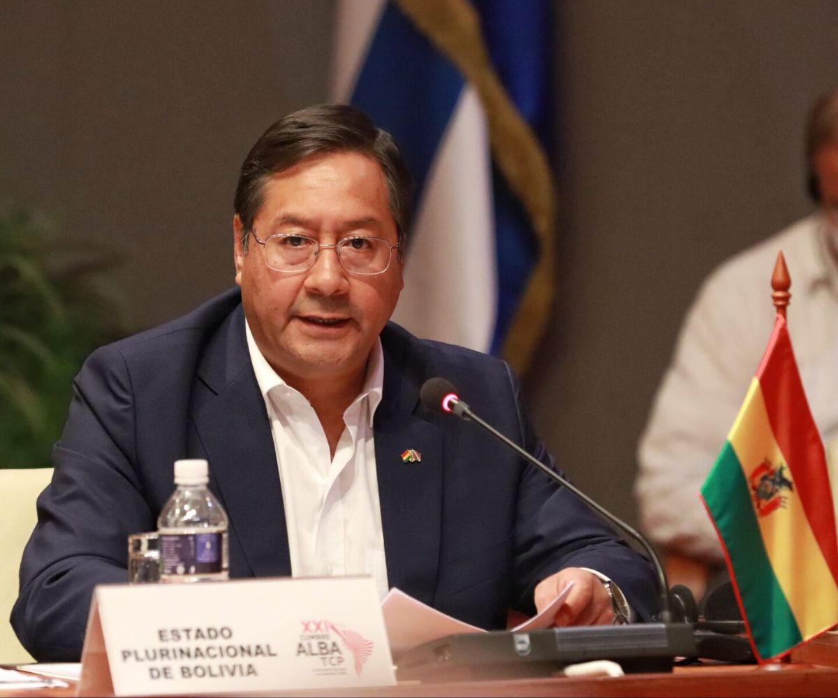 The image shows a man seated at a conference table, speaking into a microphone. He is wearing a dark navy suit with a white shirt underneath. In front of him, there is a card with text, as well as a bottle of water. A small Bolivian flag is positioned on the table. The background features a blurred flag with blue and white stripes, and greenery is visible on the left side.