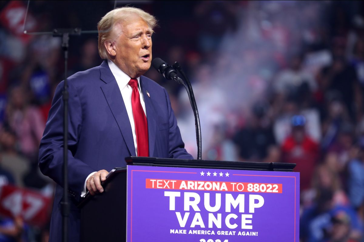 The image features a man with light hair standing at a podium, speaking into multiple microphones. He is wearing a dark blue suit with a white shirt and a red tie. The background is blurred, filled with an audience whose details are indistinct. The podium is purple and red, with text on the front.