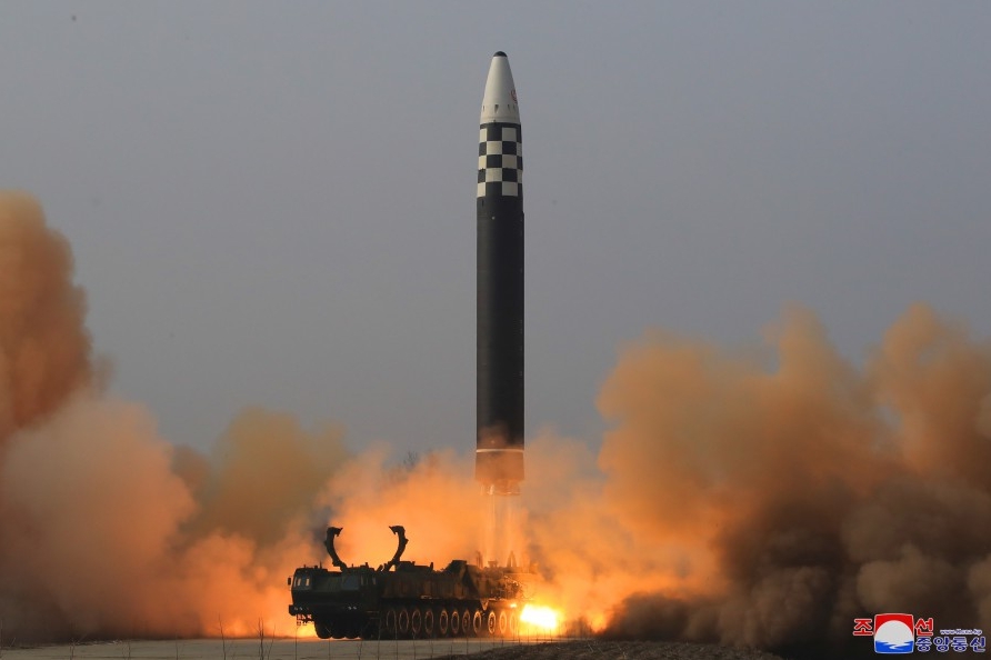 The image shows a missile launch scene. A large, black missile with a white tip and a checkered design near its upper part is ascending vertically into the sky. The background is a hazy gray, suggesting cloudy or smoky conditions. Below the missile, intense orange and yellow flames and thick gray smoke billow from the base, indicating ignition and propulsion. The missile is mounted on a massive transport vehicle with several wheels visible at the bottom. The foreground is a blurred surface, possibly the ground or launch pad. In the lower right corner, there are logos and some text in a non-Latin script.