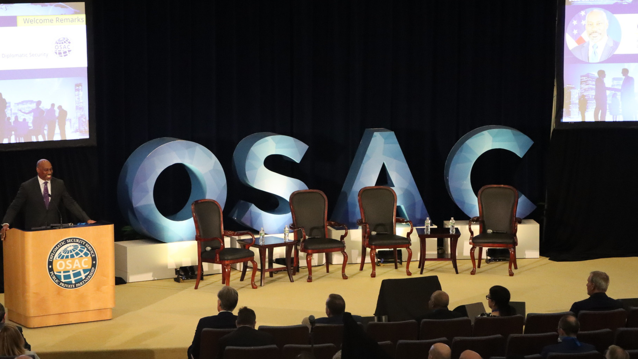 A man speaks at a podium with the OSAC logo on it, next to large letters spelling out O-S-A-C.