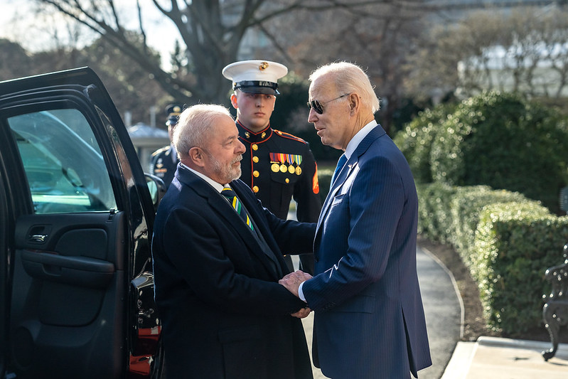 The image shows two men in formal attire standing outside near a vehicle. One man, wearing a dark suit with a white shirt and striped tie, is standing near the open door of a black SUV. He has light skin and gray hair. The other man, wearing a blue pinstripe suit and sunglasses, is facing him with his hand on the first man's arm. He also has light skin and short white hair. In the background, a member of the military, wearing a decorated uniform with a hat, stands at attention. Behind them are leafless trees and neatly trimmed hedges. The scene appears to be taking place on a bright, sunny day.