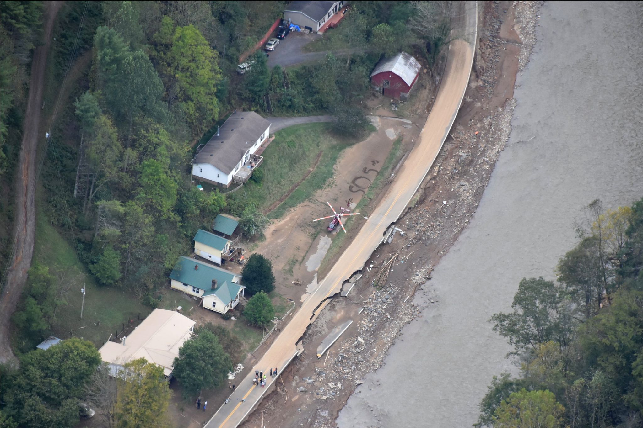 The image is an aerial view showing a small settlement adjacent to a winding road and a river. The top left section features dense green trees alongside a narrow dirt road. Nestled among the trees are several houses with different roof colors, including white, brown, and green. A red helicopter is positioned on an open space near a house with a brown roof. Adjacent to the road, the word "SOS" is marked on the ground. The road runs parallel to a river, which occupies the right side of the image. The road appears damaged near the river, with visible cracks and debris scattered along the riverbank. A group of people is gathered on the road close to the bottom of the image.