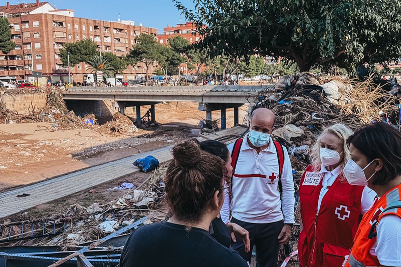 The image shows a group of people, some wearing Red Cross uniforms, at the site of a natural disaster. In the foreground, four individuals are engaged in conversation, three wearing Red Cross vests and masks. They appear concerned and attentive. One person's back is to the camera, revealing only the hair. Behind them, a large pile of debris and vegetation sits against a tree, evidence of the flood's aftermath. In the background, a muddy creek runs beneath a concrete bridge, bordered by a damaged walkway. Residential buildings and green trees are visible in the distance, under a clear blue sky.