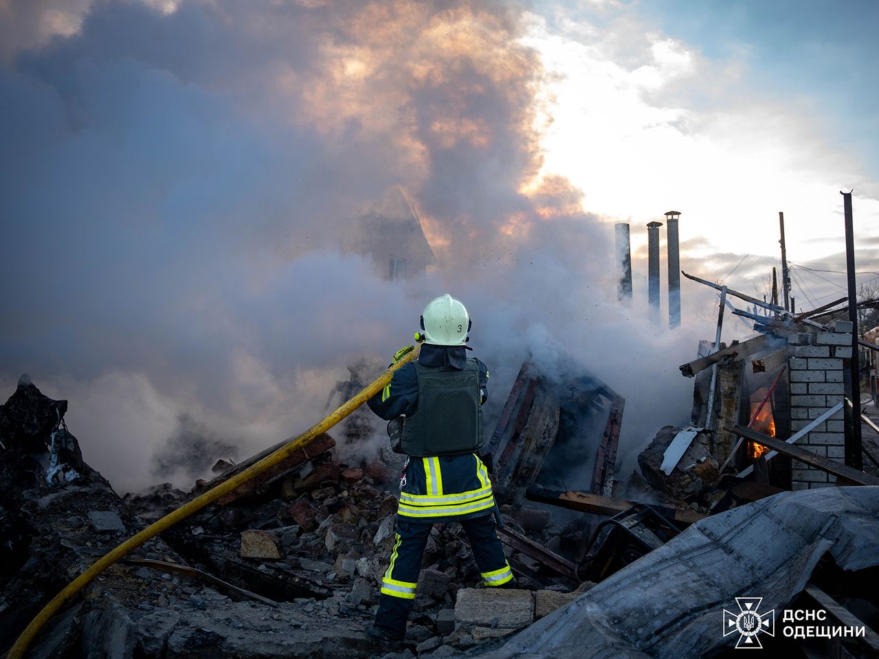 The image depicts a firefighter in protective gear, standing amidst a scene of destruction and debris. The firefighter is facing away from the camera, directing a yellow hose toward thick clouds of smoke billowing from a smoldering structure. The smoke, dense and gray, fills most of the upper portion of the image, blending into a partly cloudy sky illuminated by the sun. Debris, including chunks of concrete and scattered wooden beams, is visible on the ground. In the background, several vertical metal poles protrude through the smoke. The bottom right corner of the image contains a logo and text.