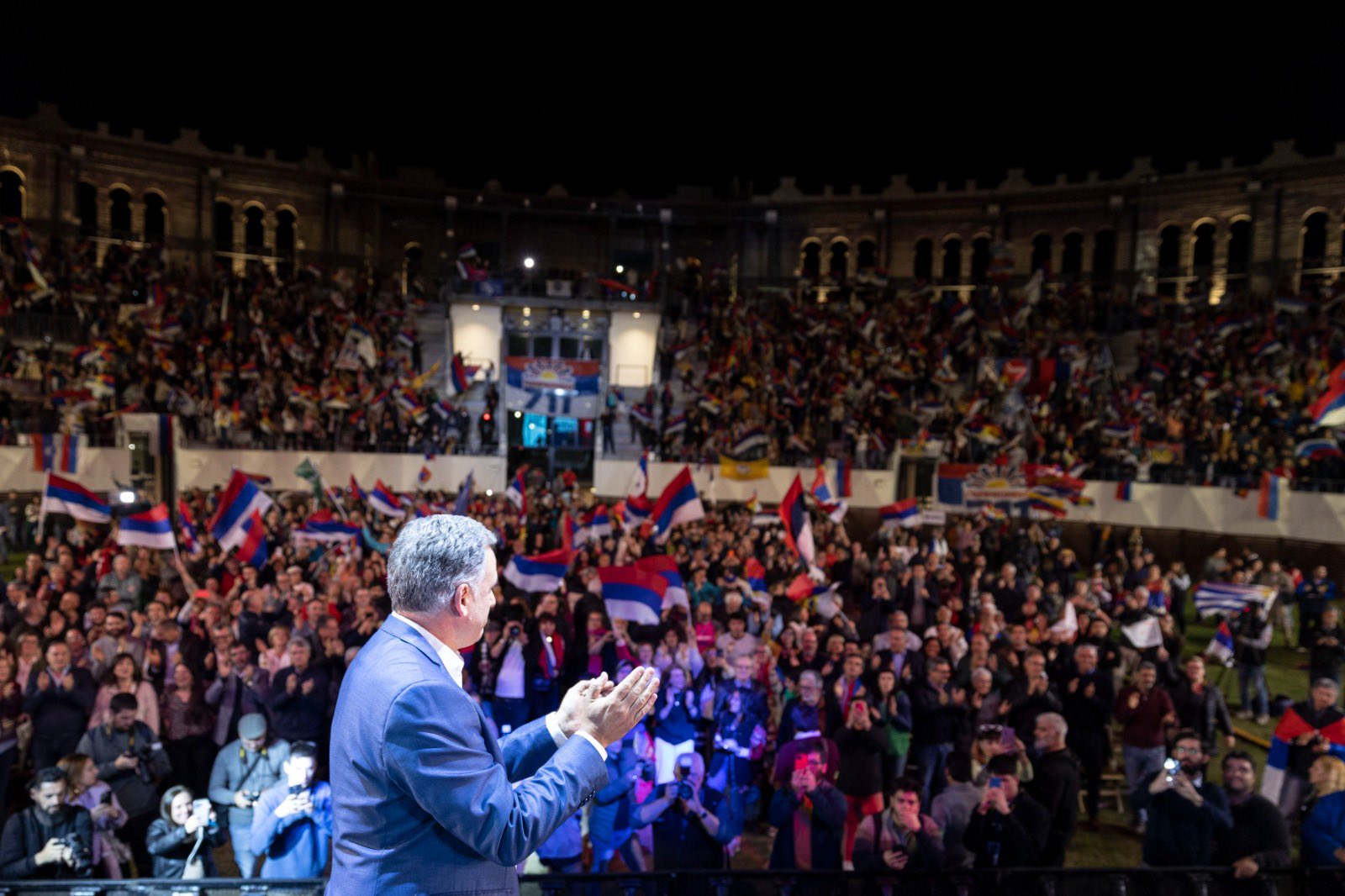 The image shows a large crowd gathered in an open-air amphitheater, illuminated against the night sky. In the foreground, a person in a suit is seen from the back, clapping while facing the crowd. The audience is densely packed, many waving red, blue, and white flags, creating a vibrant and dynamic scene. The amphitheater is grand, with rows of arches in the background. The mood is lively, and the crowd appears engaged and celebratory.