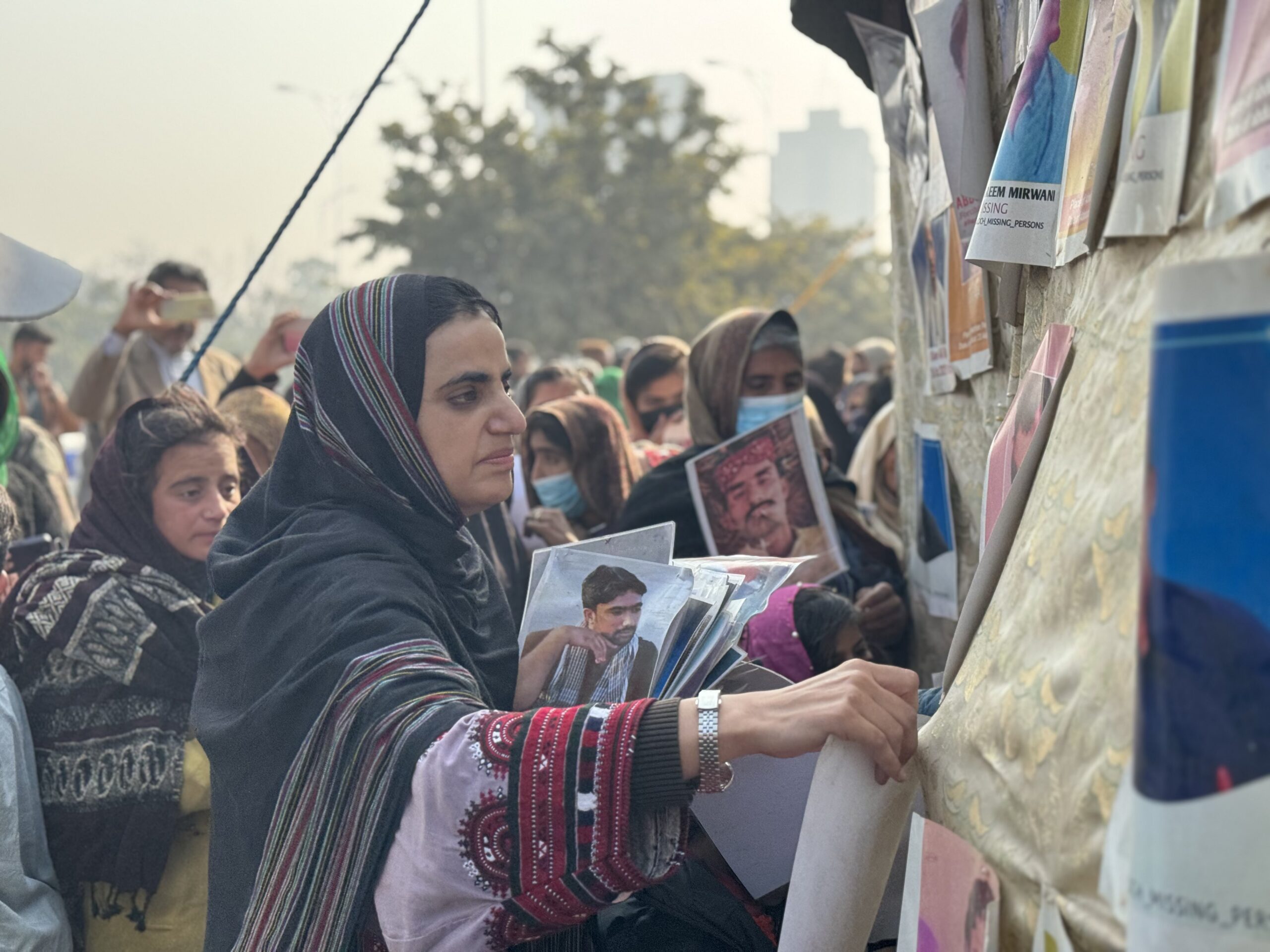 The image shows a group of people gathered, many holding photographs, possibly in a protest or memorial event. The focus is on a woman in the foreground, wearing a black shawl with colorful stripes, as she hangs a photo on a fabric-covered surface. This surface is filled with various images and papers, some of which have partially visible text suggesting missing persons. The background is slightly blurred, featuring more individuals, some wearing masks, standing amidst trees and urban structures under a hazy sky.