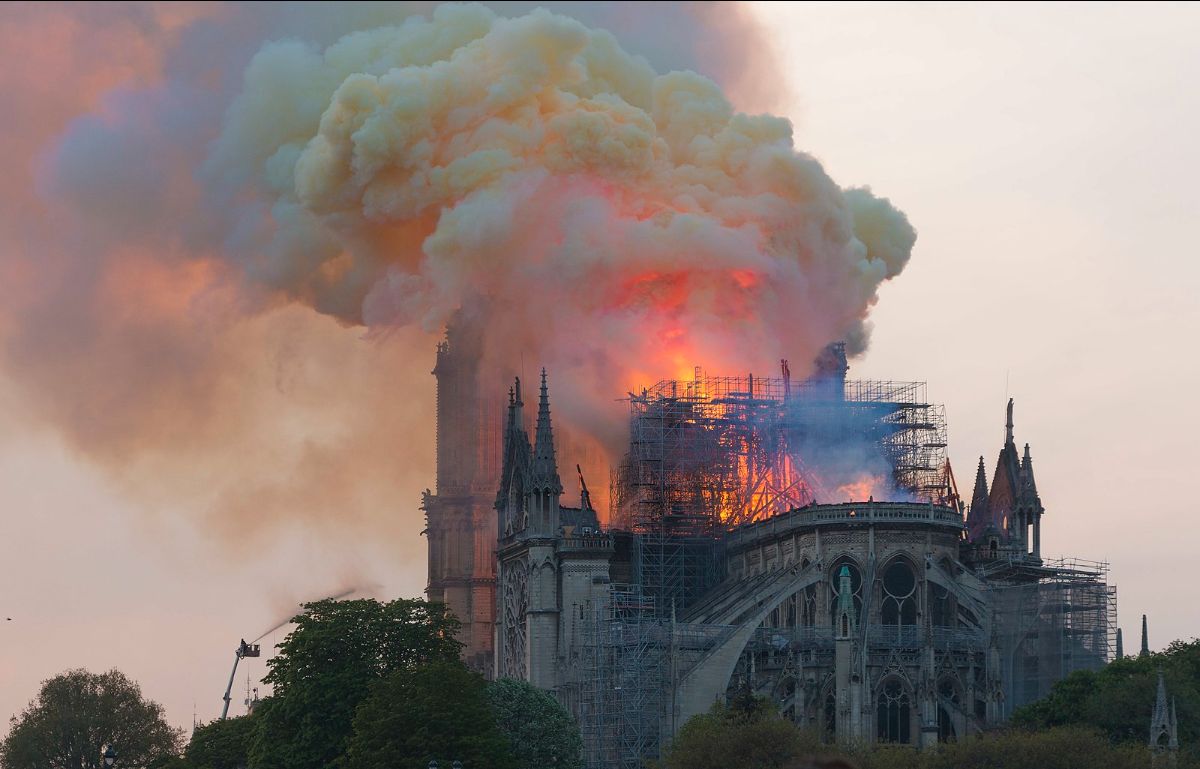 The image displays a dramatic scene of a cathedral engulfed in flames. Large, billowing clouds of orange and gray smoke rise into the sky from the top of the historical building. The fire is consuming the roof, with visible bright flames and intense red and orange hues. Parts of the cathedral are surrounded by scaffolding, indicating ongoing construction or renovation. The intricate gothic architecture of the cathedral is visible, with pointed arches and ornate detailing. In the foreground, there are trees and a silhouetted crane, possibly used by firefighters to manage the blaze.