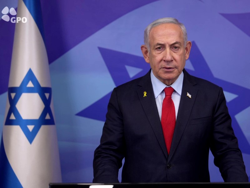 Photo of a man in a suit with a red tie standing beside the flag of Israel.