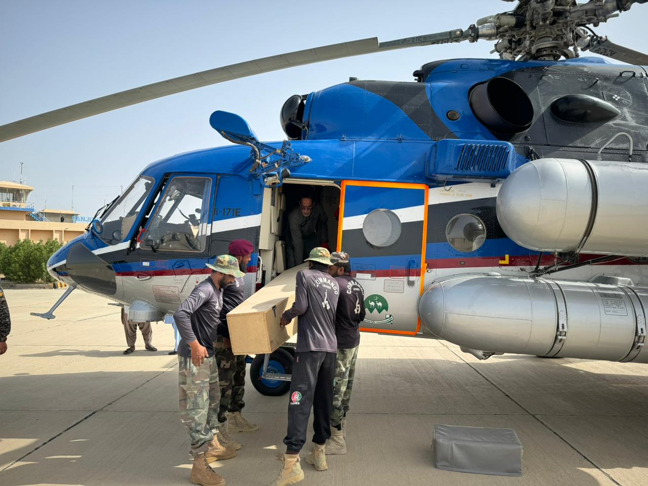 The image shows a group of uniformed men unloading a large cardboard box from a blue and white helicopter on a concrete tarmac. The helicopter, a Mi-17E, is parked with its door open, revealing the interior where one person is visible. The men, wearing camouflage pants and shirts with logos, work together to carefully lift the box. The helicopter is equipped with large cylindrical fuel tanks on its sides and a rotor is visible at the top. In the background, a beige building and some greenery are seen under a clear blue sky.