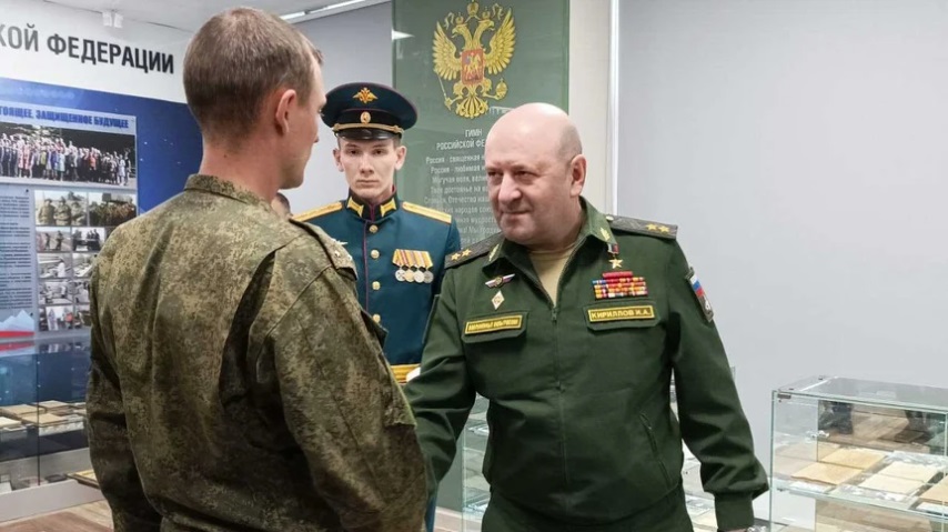 The image shows three men in military uniforms inside a room with display cases. The man in the foreground, seen from behind, is wearing a green, camouflage-patterned military uniform. The man in the center and to the right is bald and smiling, wearing a green military uniform with various patches and insignia. His uniform features a name tag, and he is shaking hands with the man in camouflage. Behind them, another man in a more formal blue uniform with medals and a cap stands, observing the interaction. The background contains a wall with information panels and a large emblem featuring a double-headed eagle, associated with Russia.