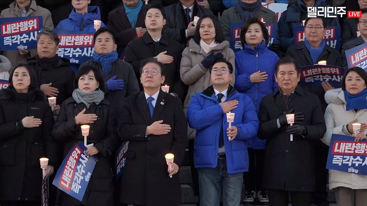 The image portrays a group of people standing closely together on a set of stairs. Each person is holding a lit candle, with most of them placing their other hand over their heart. They are dressed in winter clothing, including coats and scarves, as the scene likely takes place during cold weather. Several individuals in the group are holding signs with Korean text. The atmosphere suggests a solemn or significant event, possibly a vigil or demonstration. The backdrop is a darkened area, hinting that the event might occur during the evening. The general mood seems serious and unified.