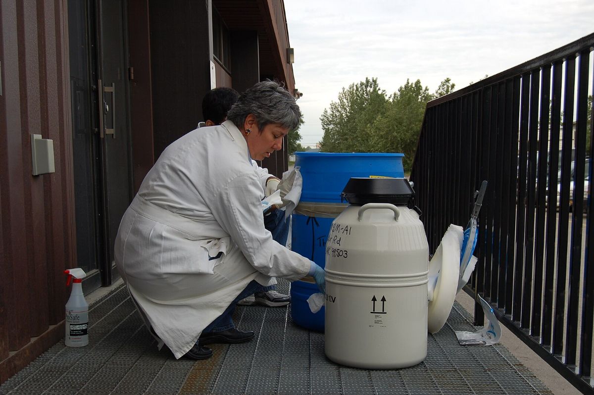 The image depicts a person wearing a white lab coat and blue gloves, kneeling on a metal grate outdoor walkway next to a building. They are interacting with large laboratory containers. In the foreground, there is a white cylindrical container with black text and arrows, next to a larger blue plastic drum. The background shows a railing and trees under a cloudy sky. The setting suggests an industrial or laboratory environment.