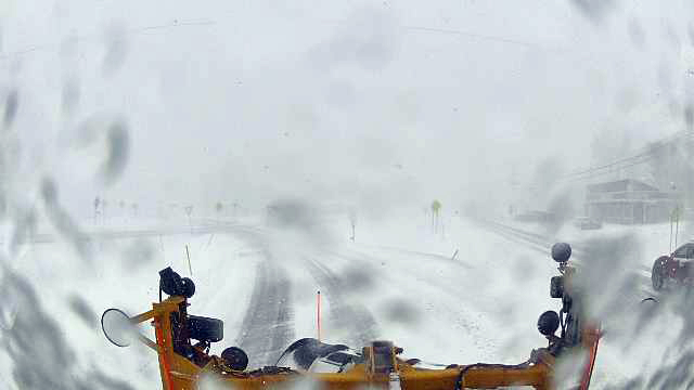 The image shows a snow-covered road viewed through the windshield of a snowplow. The snowplow's yellow frame is visible at the bottom of the image, with several black and white mechanical parts and lights. Snow heavily covers the road and the road signs are barely visible due to the dense snowfall. The sky is overcast, blending into the white background, and the visibility is low.