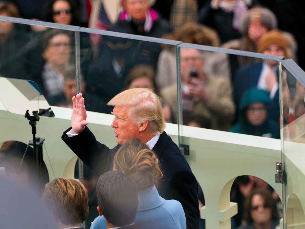 The image shows a public event with a man at the center, who appears to be making a gesture with his right hand raised. He is wearing a dark overcoat and has light blonde hair. The event takes place in an outdoor setting with a partially visible glass barrier around the speaker. In the foreground, there are a few individuals with their backs to the camera, with one partially obscured by the man’s broad shoulders. Behind them, a crowd of people can be seen, some taking photos or videos and others simply watching. Several people in the crowd are wearing winter clothing, including scarves and hats, hinting at a cold day. The setting appears formal, possibly ceremonial, given the arrangement and attention of the attendees.