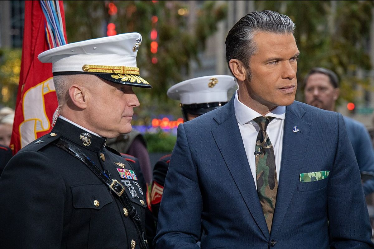 The image shows two men standing side by side. On the left, a man in a Marine Corps dress uniform with a white peaked cap decorated with gold and black details stands in profile. His uniform is adorned with several medals and colorful ribbons. A red Marine flag with gold trim is partially visible behind him. On the right, a man in a blue suit with a camouflage-patterned tie and pocket square looks slightly to his left. He has neatly styled hair and is facing away from the camera. The background is blurred, featuring greenery and soft, colorful lights, suggesting an outdoor setting, possibly a public event or ceremony.