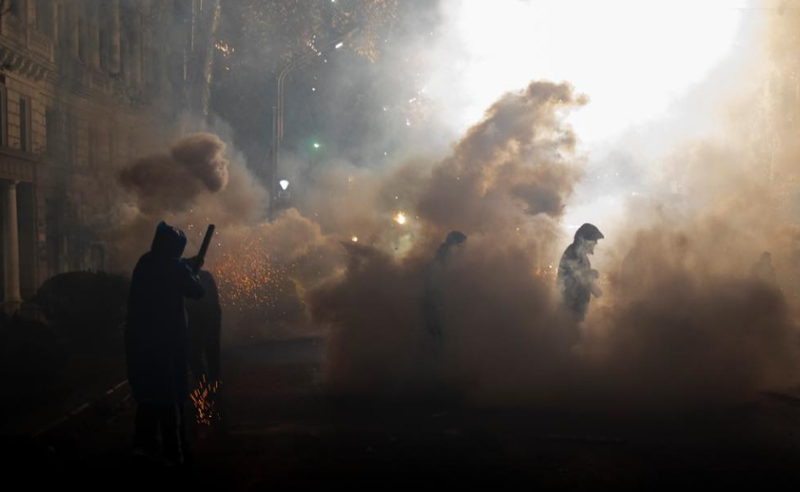 The image depicts a scene at nighttime, with several figures obscured by large clouds of smoke on a city street. The smoke is illuminated by bright lights from above, creating a dramatic contrast against the dark surroundings. Silhouetted figures, some holding objects resembling batons, are partially visible amidst the thick smoke. The left side of the image shows the edge of a building with classical architectural details, faintly illuminated by ambient light. Sparks or small glowing particles are visible through the smoke, adding to the intensity of the scene.