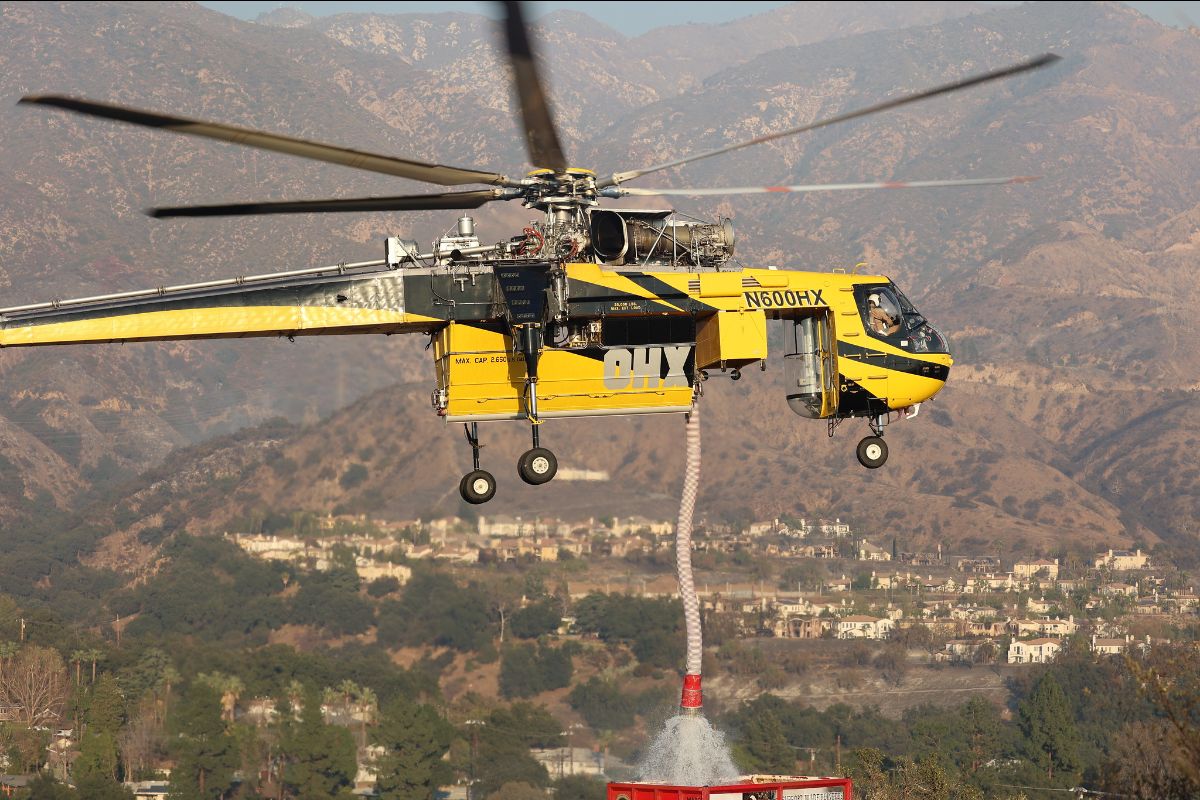 The image depicts a yellow and black firefighting helicopter, model number N600HX, in flight against a mountainous backdrop. The helicopter's large rotor blades are spinning, and its landing gear is visible beneath it. A long hose, attached to the underside of the helicopter, is releasing water over a target below. The background features rugged hills and a residential area with scattered houses and green vegetation.