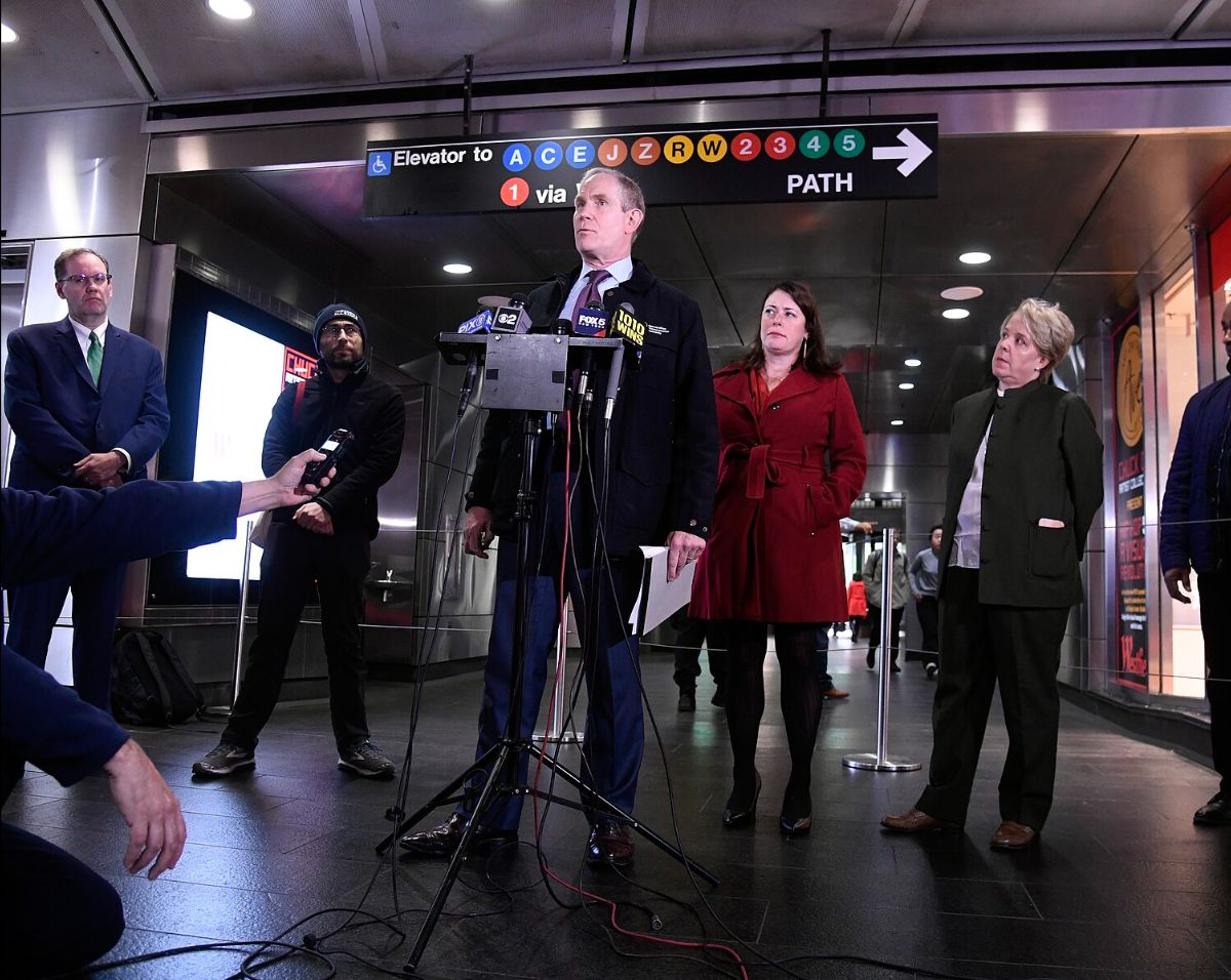 The image shows a group of six people standing in a subway station during a press conference. The central figure, dressed in a dark suit and tie, is speaking at a podium equipped with multiple microphones from various media outlets. The people are positioned indoors under a ceiling with circular lights, and the floor appears to be dark, glossy tile. Behind the group, a sign displays directions for elevators to subway lines A, C, E, J, Z, R, W, and numbers 2, 3, 4, and 5, along with an arrow pointing to the right for PATH. The background features stainless steel walls and digital signage, enhancing the modern aesthetic of the subway station.