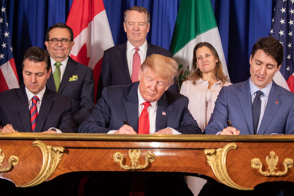 The image depicts three men seated at an ornately carved wooden table, each signing documents. The man on the left is wearing a dark suit with a red striped tie, the man in the middle is wearing a dark suit with a red tie, and the man on the right is wearing a blue suit with a dark tie. Behind them, two men and one woman stand, all in business attire, smiling. The background shows the flags of the United States, Canada, and Mexico.