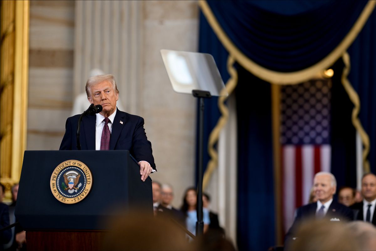 The image depicts a formal event set in an opulent room characterized by marble walls and draped blue curtains with gold trim. A man is standing behind a podium adorned with the seal of the President of the United States. He is speaking into a microphone. The podium is positioned slightly to the left of the image. Directly behind him is a blurred teleprompter screen. In the background, a large United States flag is visible, partially obscured by the people in attendance. The attendees, who are out of focus, sit attentively in rows.