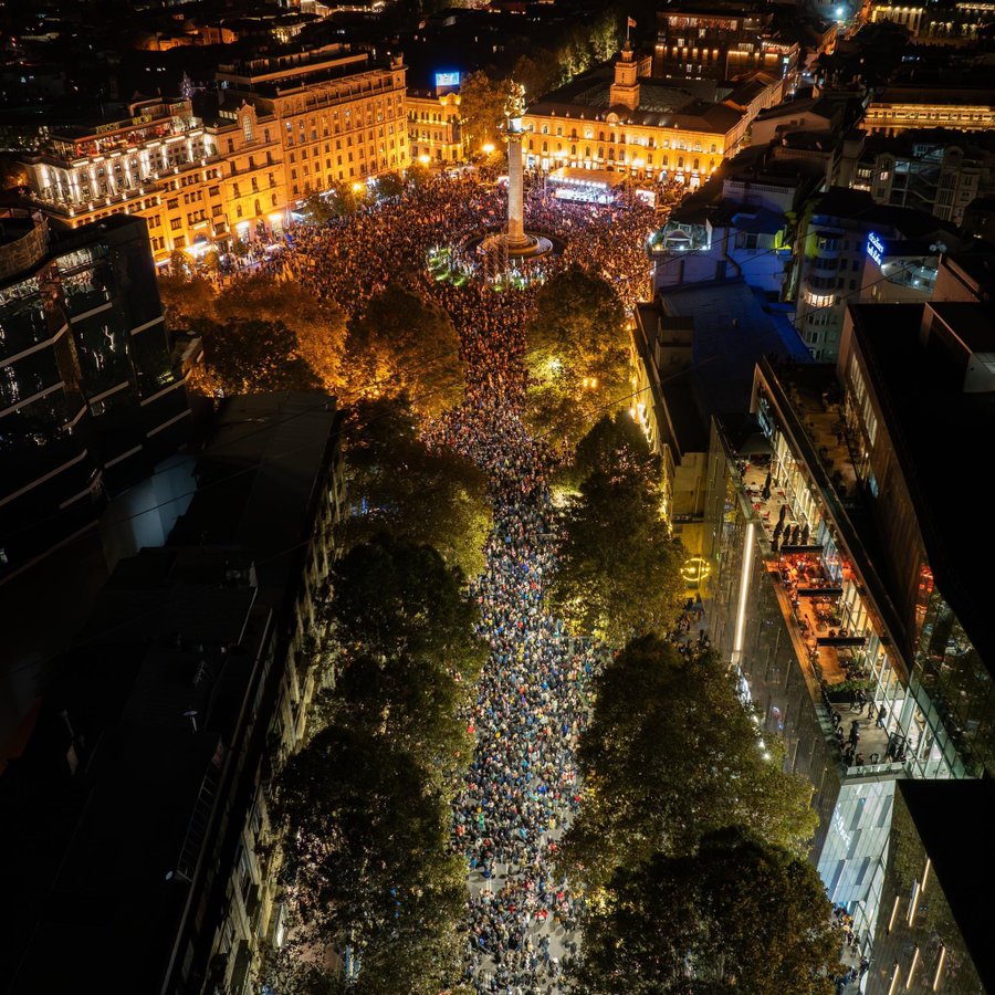 The image is an aerial view of a large, brightly lit urban plaza at night, teeming with a massive crowd of people. The scene is dominated by a central column-like monument surrounded by a roundabout, where the crowd appears to be most concentrated. Surrounding the plaza are various buildings, some of which are illuminated with decorative lights, casting a warm glow over the area. The roads leading away from the plaza are filled with people, indicating a gathering or event. On the right, a glass-fronted building with visible floors and people inside indicates a bustling city atmosphere. Trees interspersed along the streets add greenery to the urban setting. The contrast between the dark sky and the vibrant lights enhances the lively ambiance of the scene.